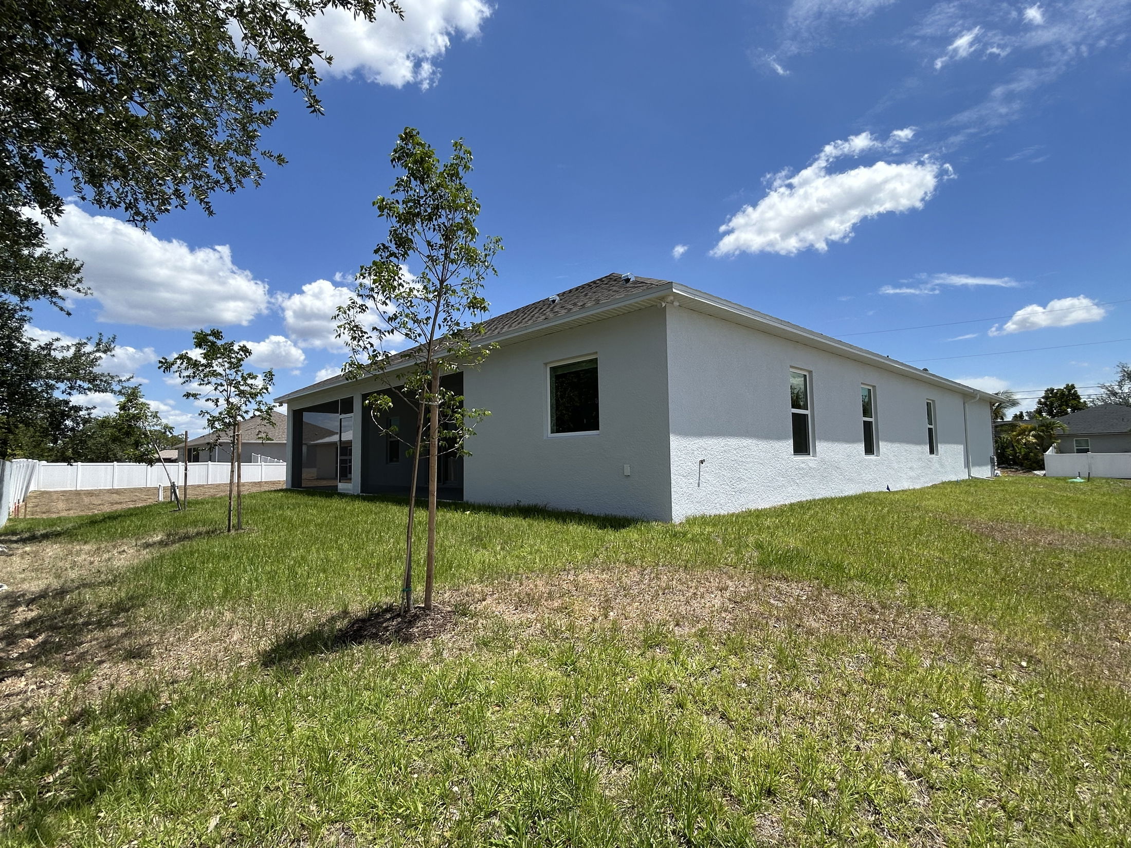 Side view of a modern, single-story white house with a spacious grassy backyard and young trees under a blue sky with fluffy clouds.