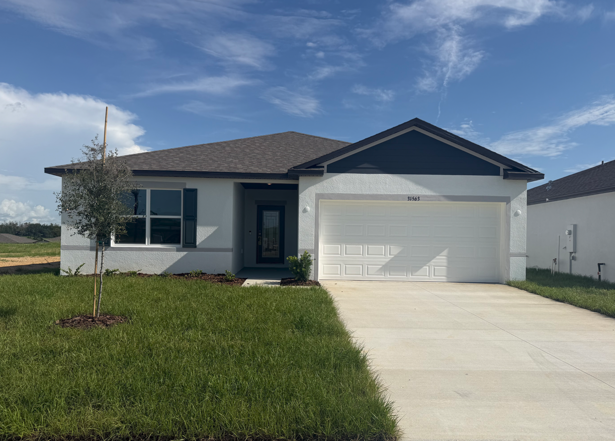 Single-story house with a light exterior, dark roof, and a two-car garage, surrounded by green grass under a blue sky.
