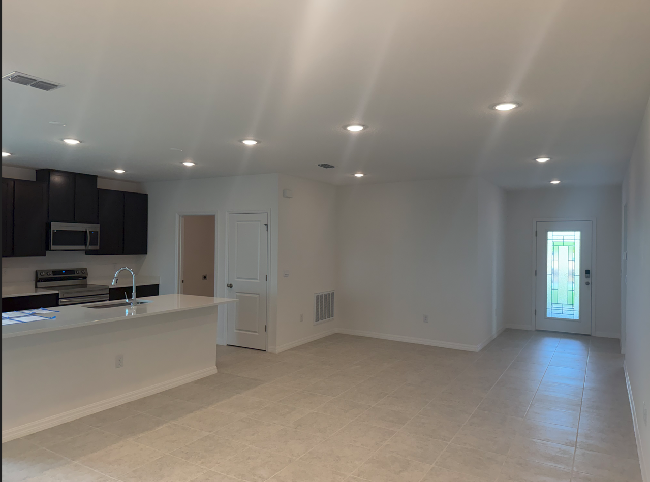 A modern, open-concept living space with tile flooring, a kitchen island, and natural light from a glass door leading outside.