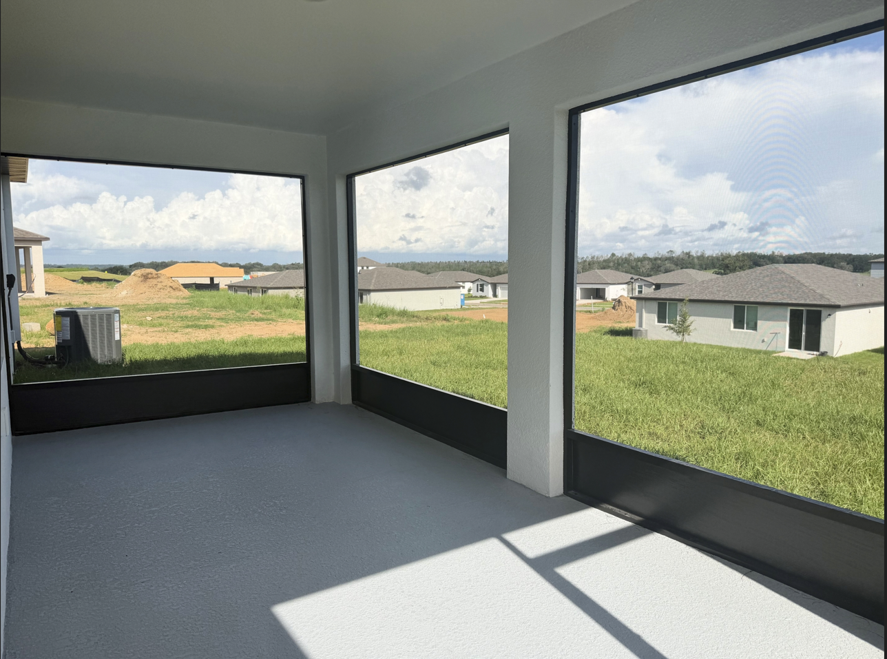 A screened porch with large windows overlooking a grassy field and nearby houses under a cloudy sky.