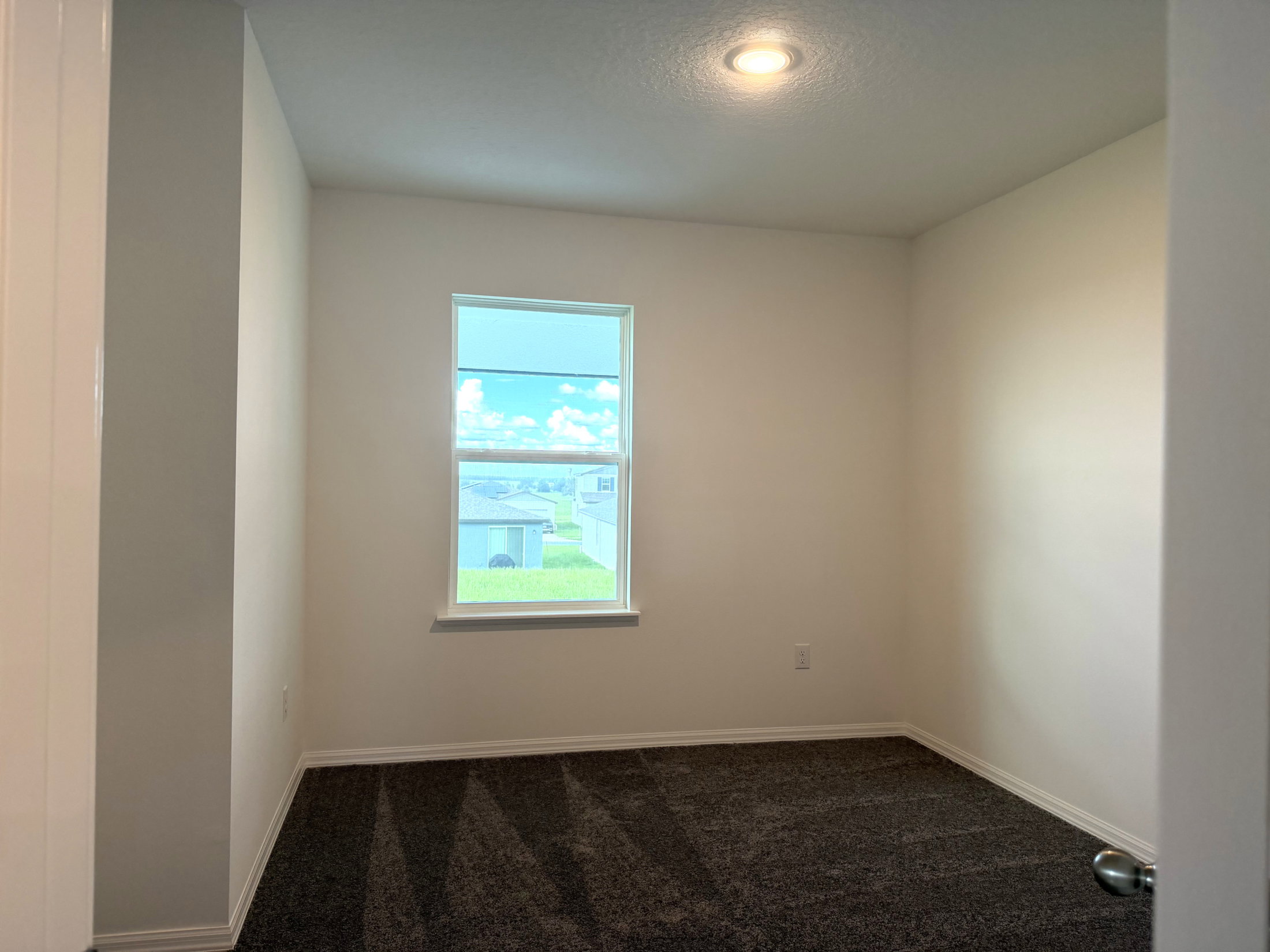 A vacant room with light-colored walls, a window showing a blue sky, and carpeted flooring.