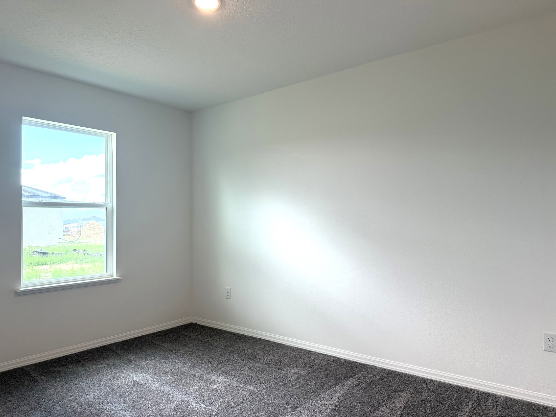 Empty room with light-colored walls and a gray carpet, featuring a window that allows natural light to enter.
