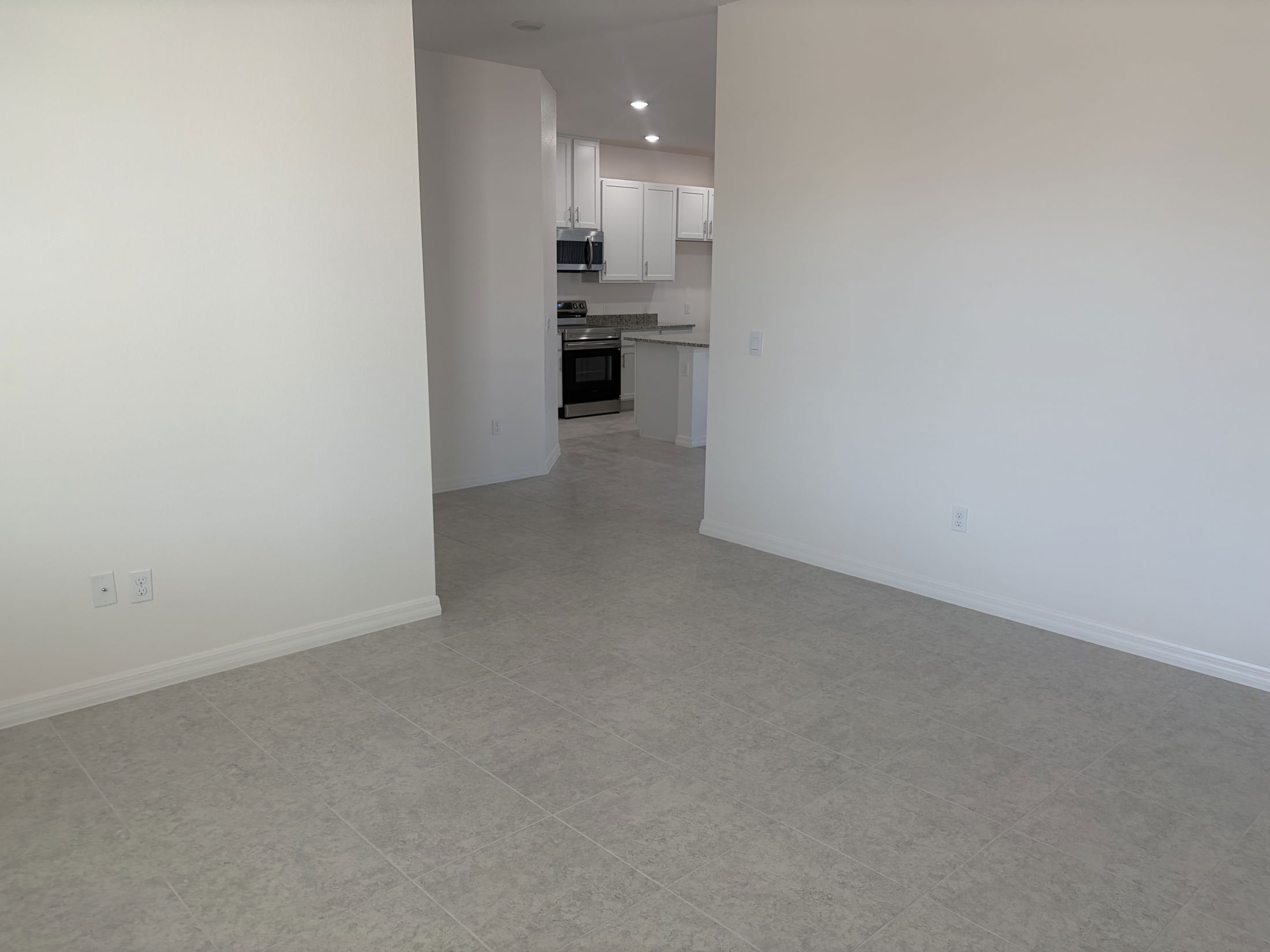 Spacious living area showcasing tiled flooring and a view into a modern kitchen with white cabinetry.