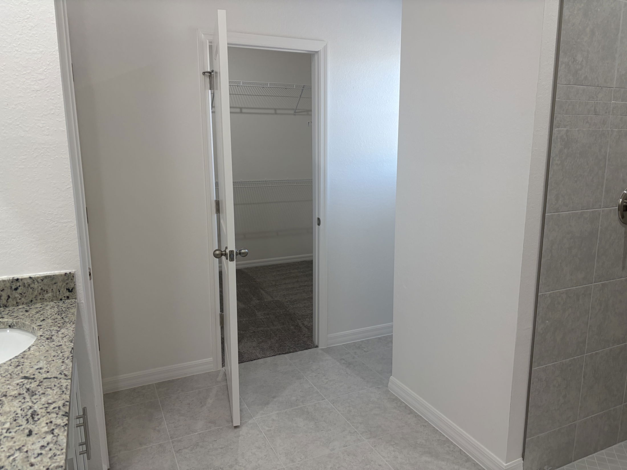 Modern bathroom interior featuring a spacious walk-in closet with an open door, designed with gray tile flooring and a granite countertop vanity.