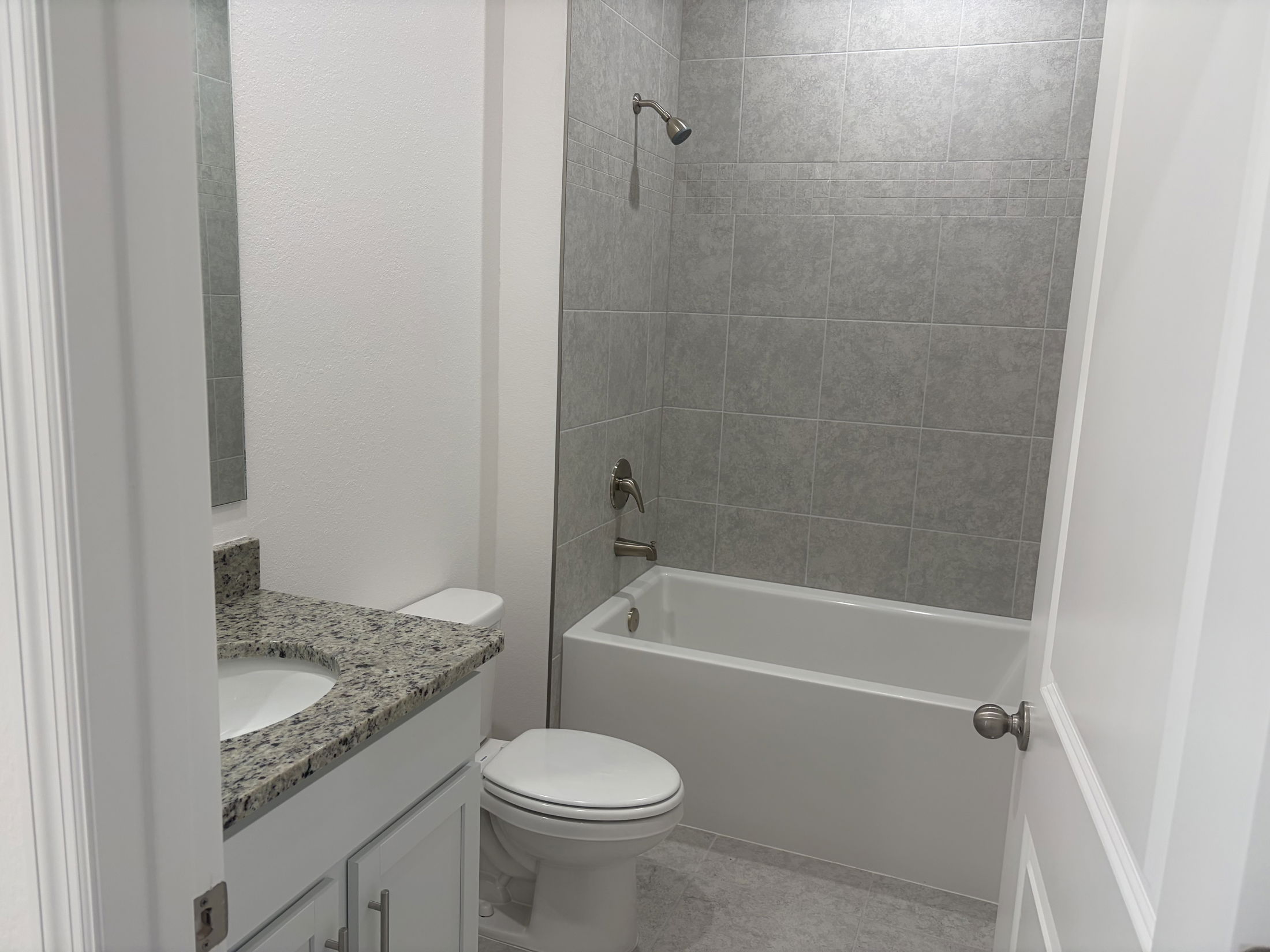 Modern bathroom featuring a grey tiled shower, white bathtub, granite countertop vanity, and sleek fixtures.