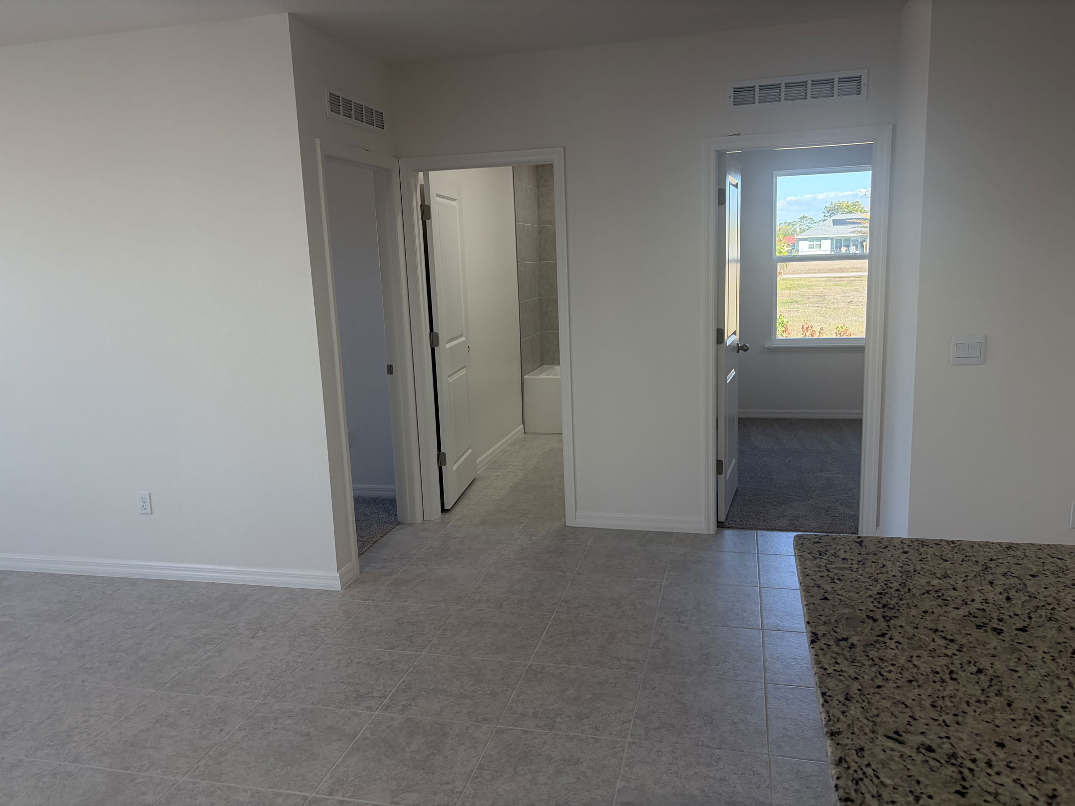 Bright interior view of a modern apartment showcasing tile flooring, doorways leading to a bathroom and a carpeted room, and natural light streaming through a window.
