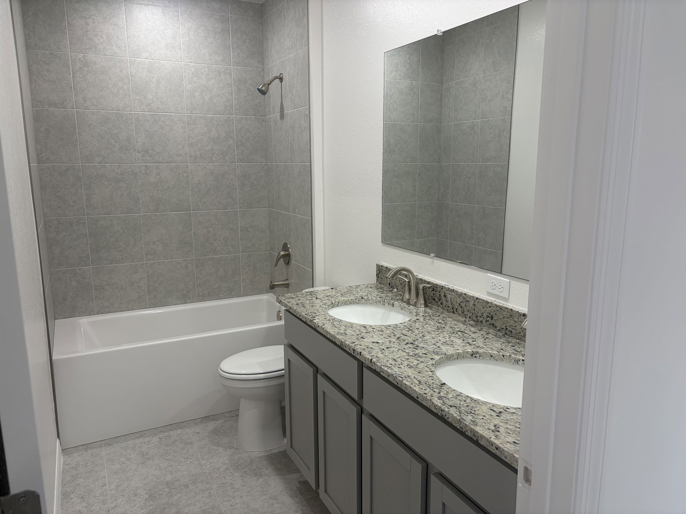 Modern bathroom featuring a double vanity sink with granite countertops, a white tub with shower, and neutral gray tile walls.