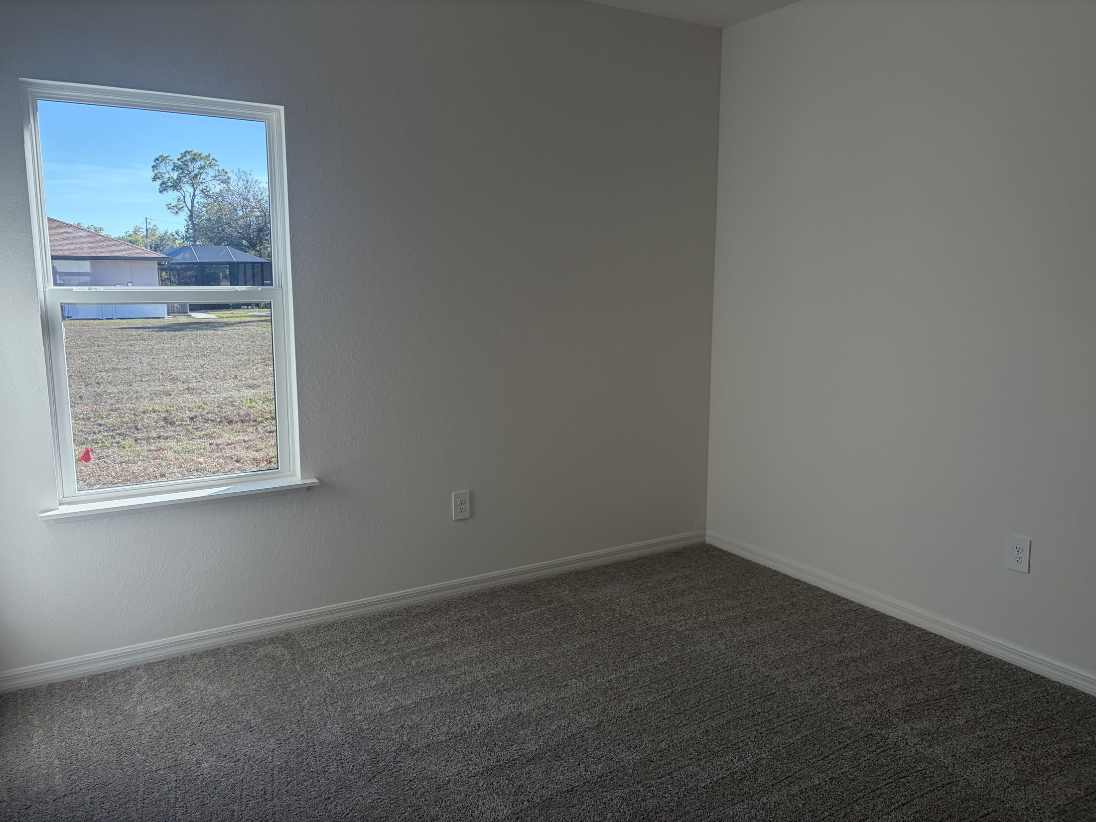 Bright and empty room with a window overlooking a grassy backyard, featuring light gray carpet and white walls.