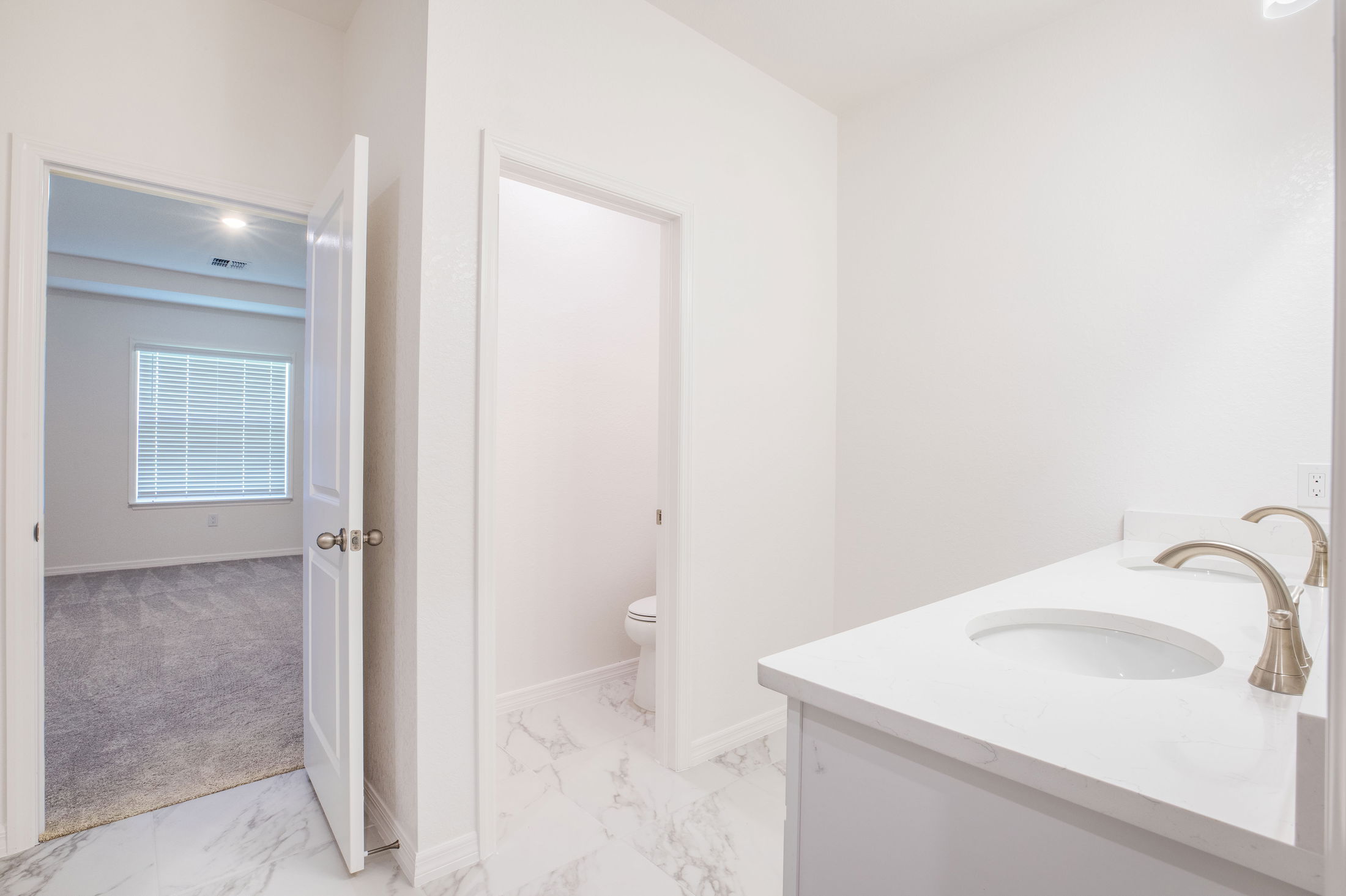 Bright, modern bathroom with marble floors, white vanity, and an open door leading to a carpeted bedroom.