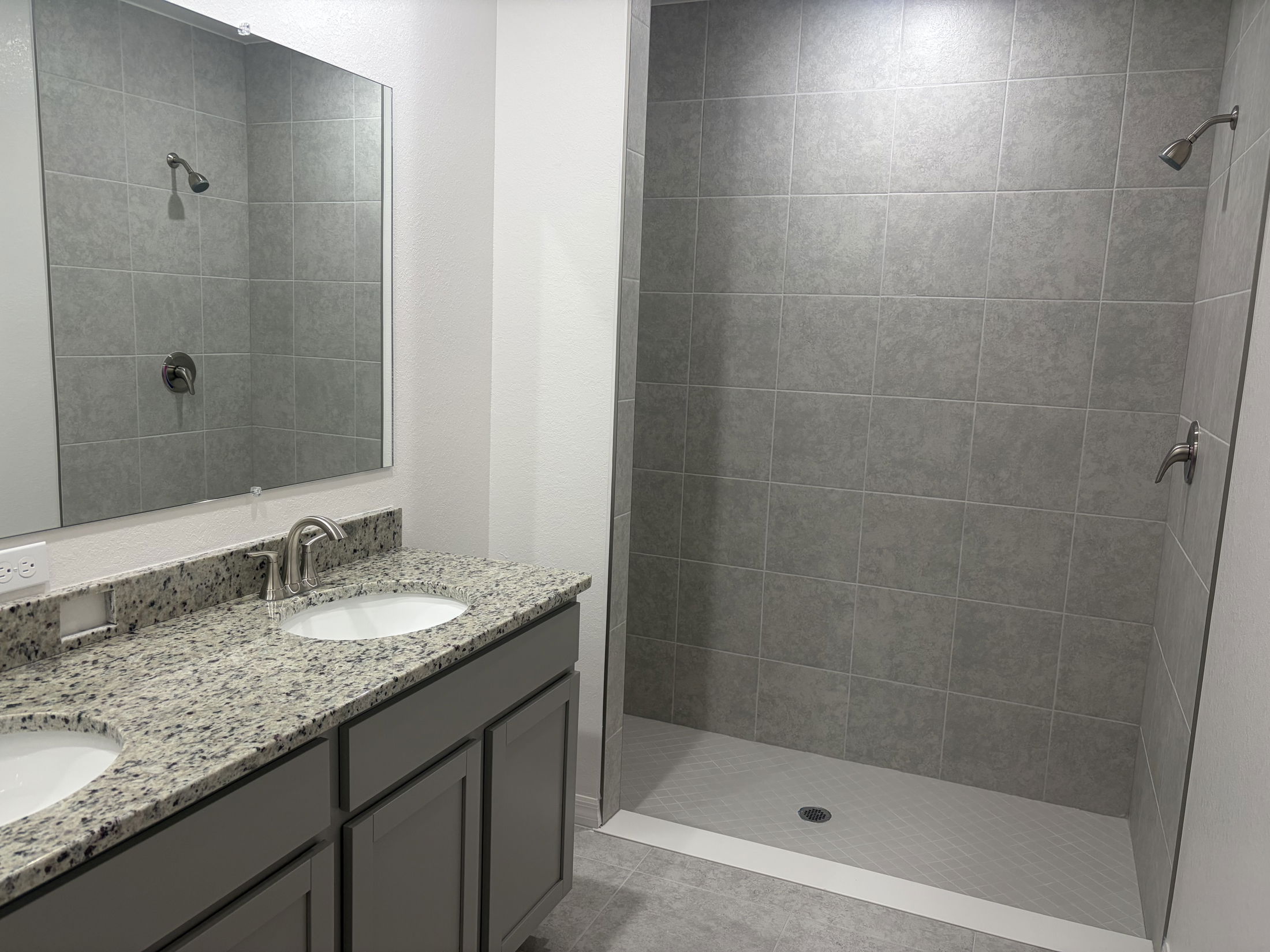 Modern bathroom featuring a double vanity with granite countertop, large mirror, and a walk-in shower with grey tiled walls.
