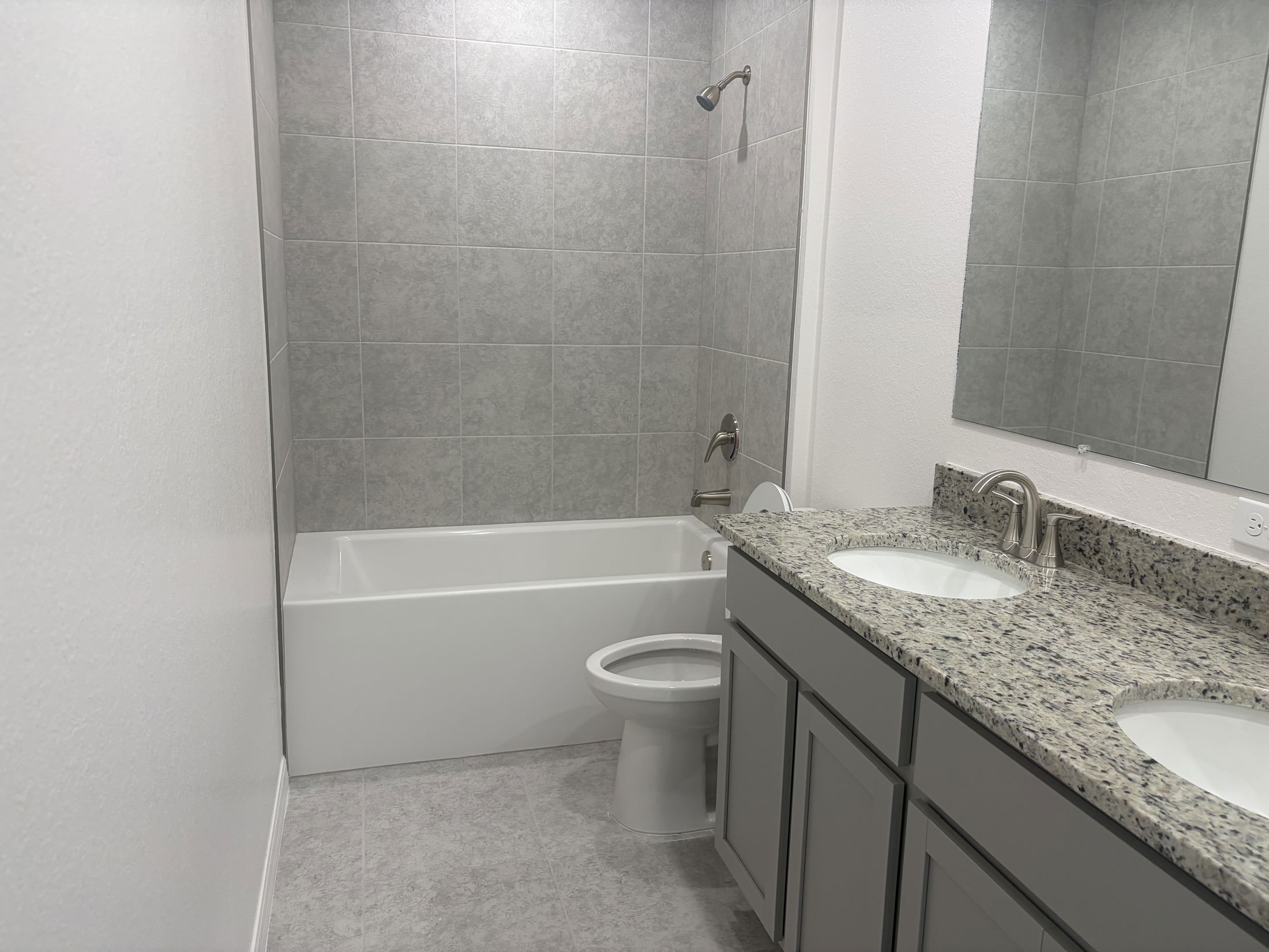 Modern bathroom featuring a gray tiled wall, a white bathtub, dual sink vanity with granite countertops, and a large mirror.