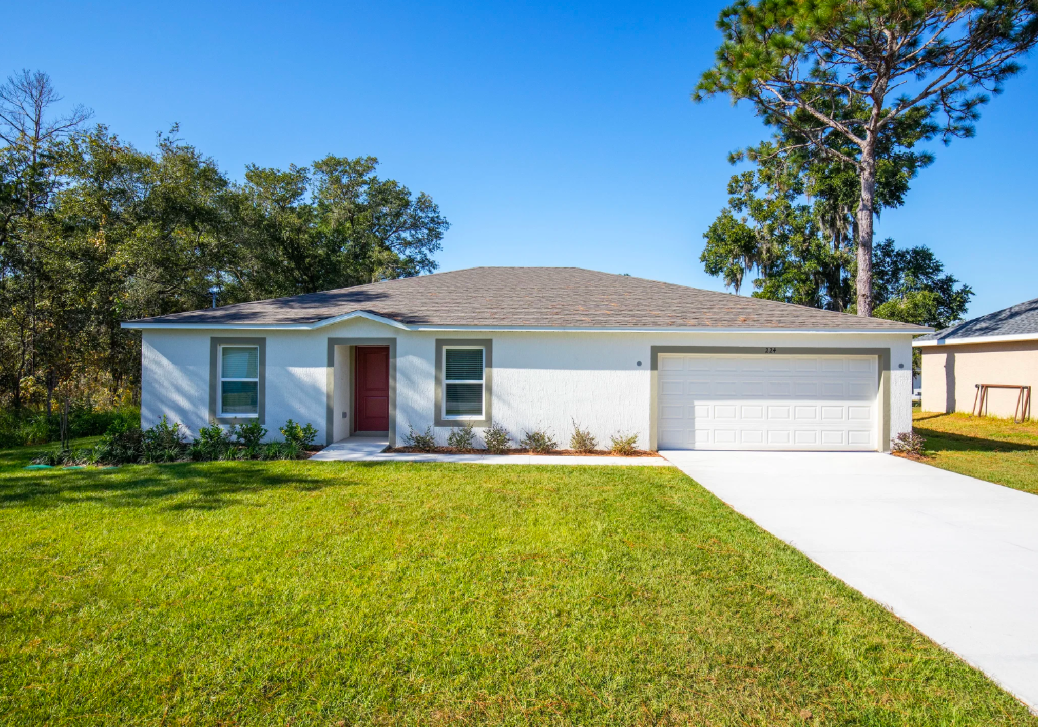Single-story white house with a red door, green lawn, and a paved driveway under a clear blue sky.