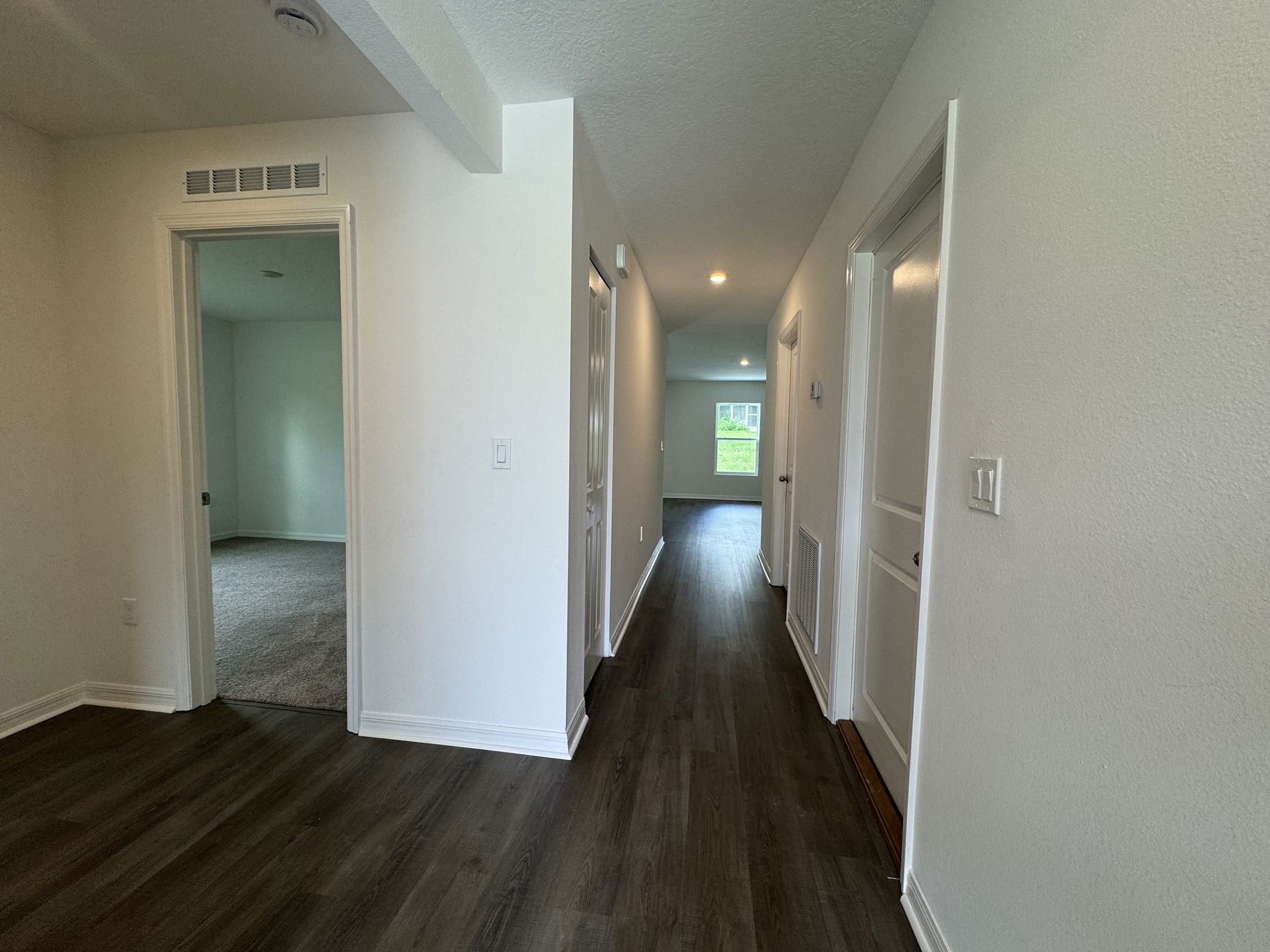 A modern corridor featuring neutral-colored walls and hardwood flooring, leading to multiple rooms and natural light at the end.