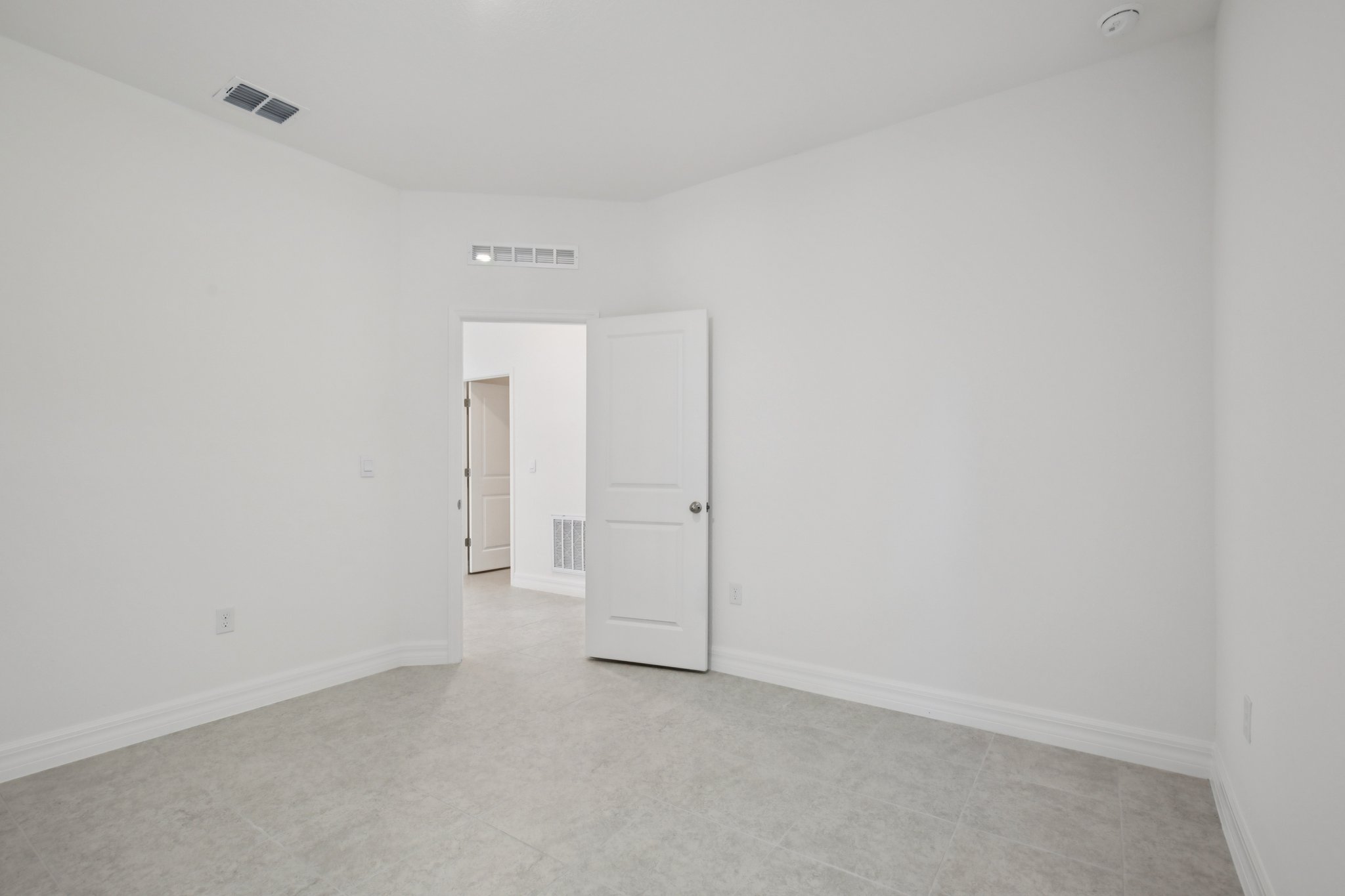 A spacious, empty room with white walls and tiled flooring, featuring an open door leading to another area.