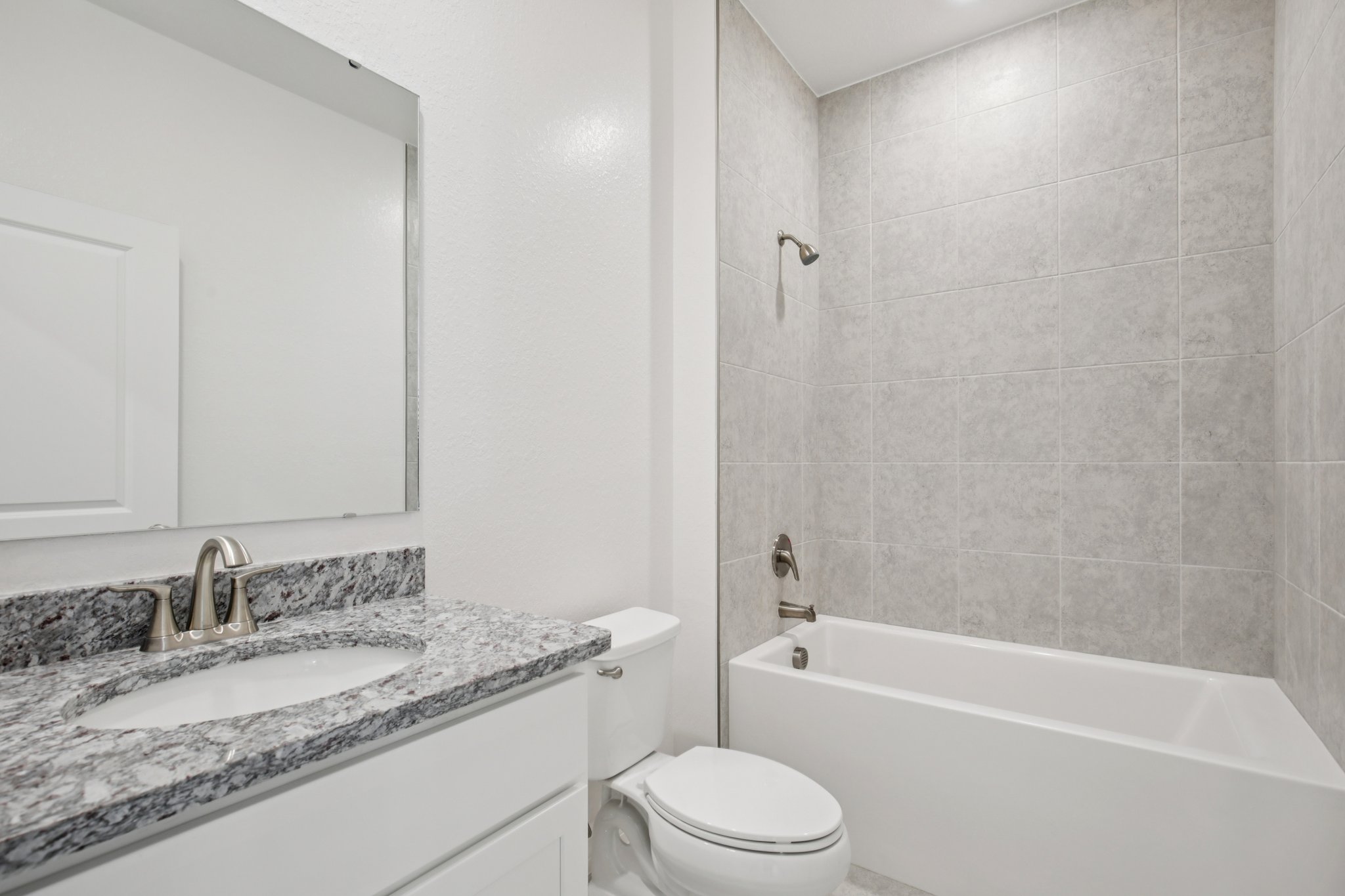 Modern bathroom interior featuring a sleek white bathtub, granite countertop, and grey tile walls with a mirror above the sink.