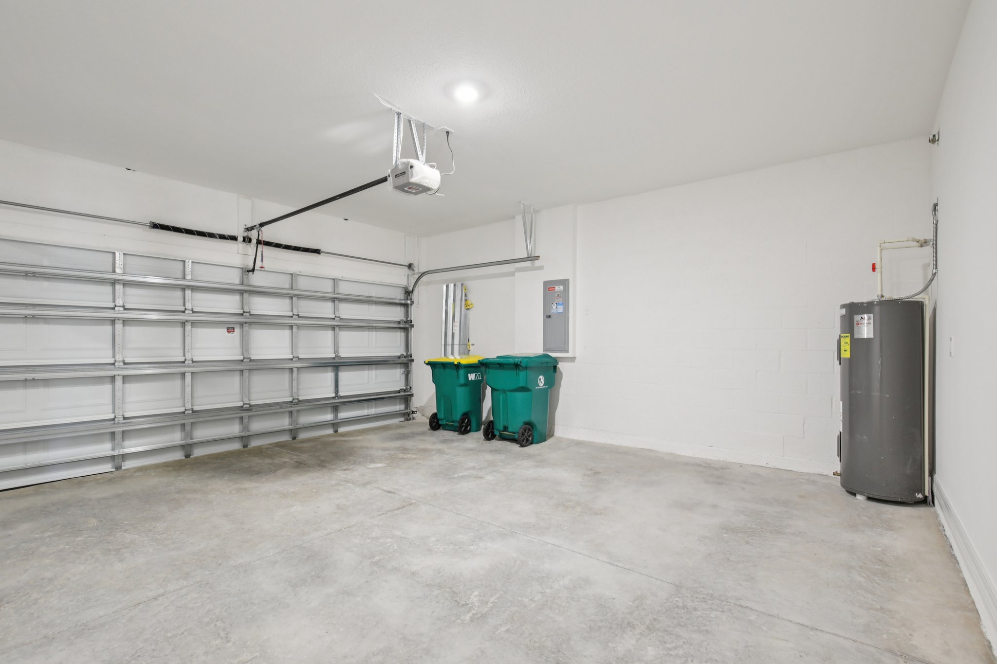 Interior view of a spacious, clean garage featuring a gray water heater and two green trash bins against a bright white wall.