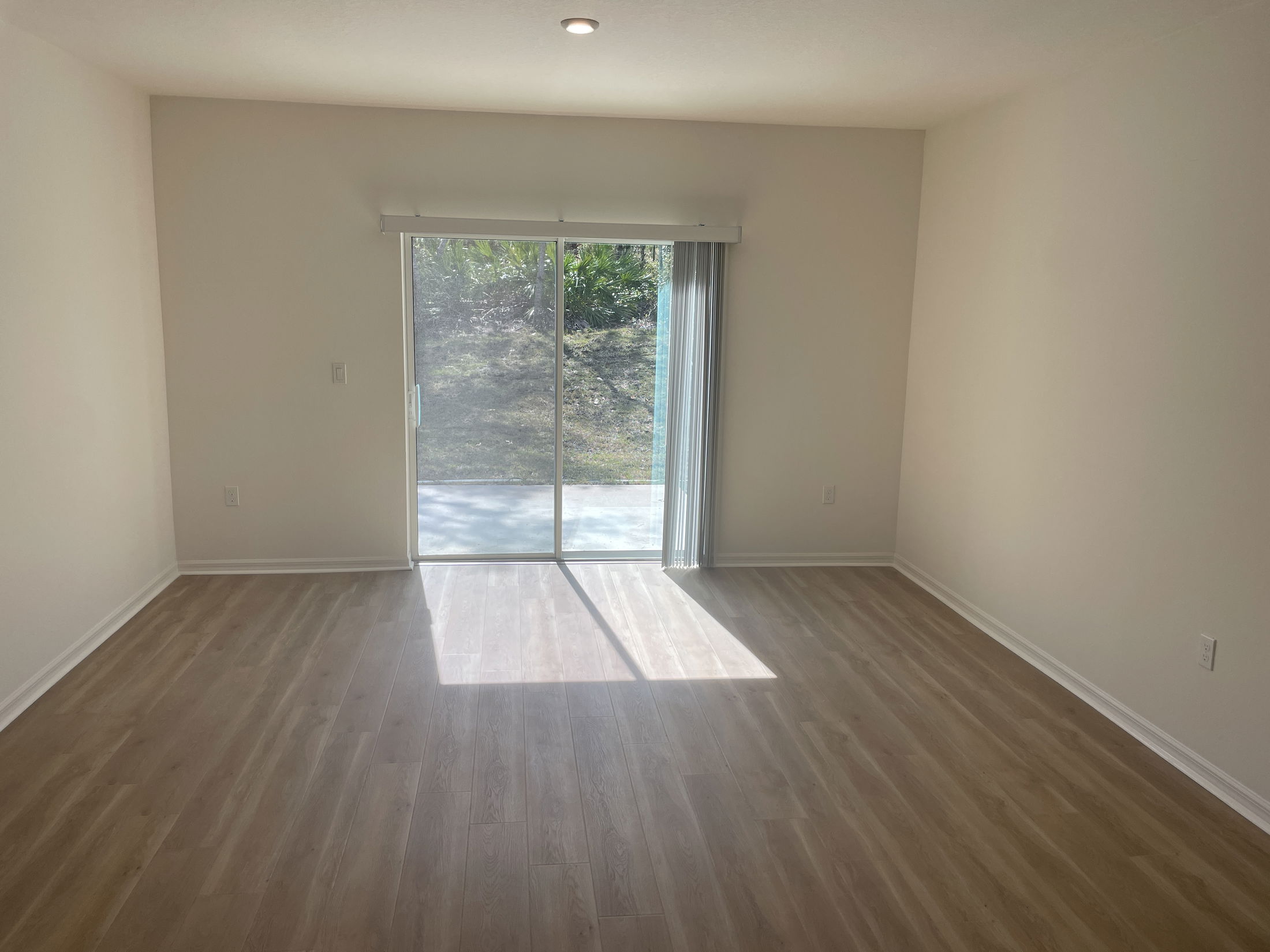 Empty room with light wooden flooring and sliding glass doors leading to a sunlit patio.