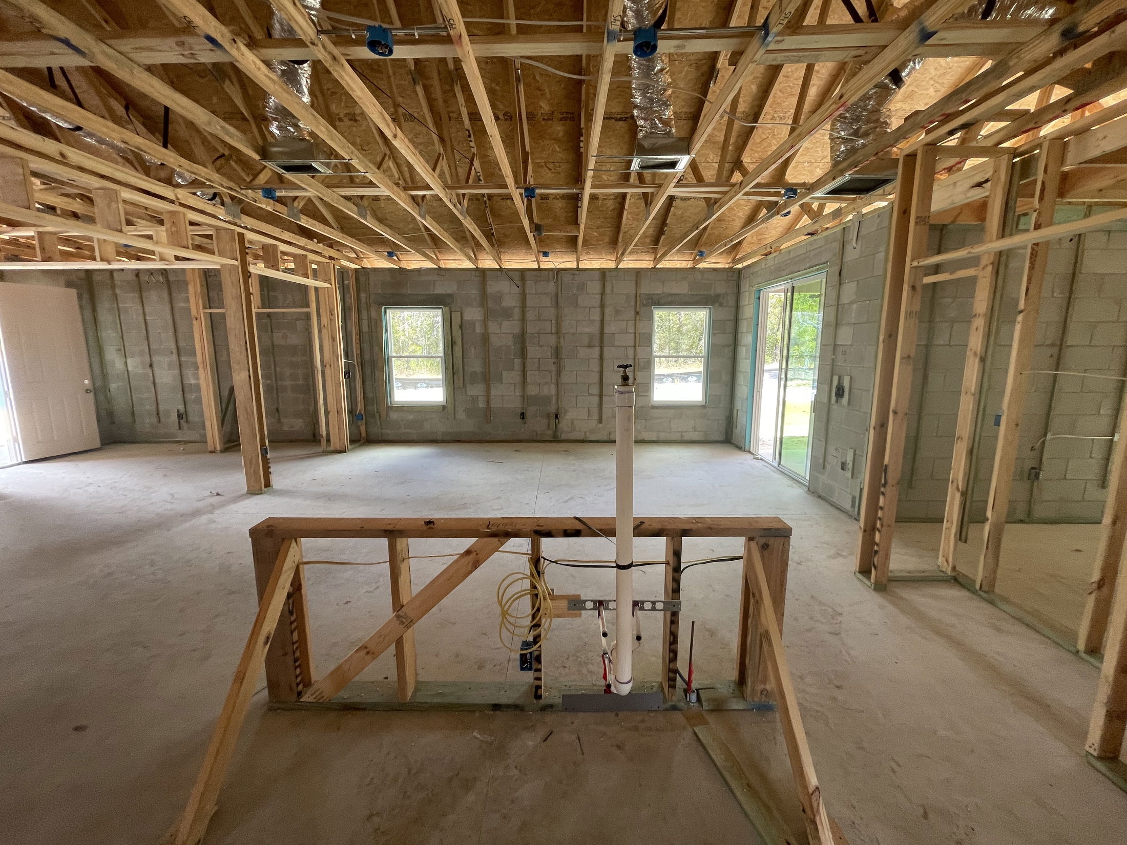 Interior view of a house under construction, showcasing exposed wooden framing and unfinished concrete walls with installed windows and sliding doors.