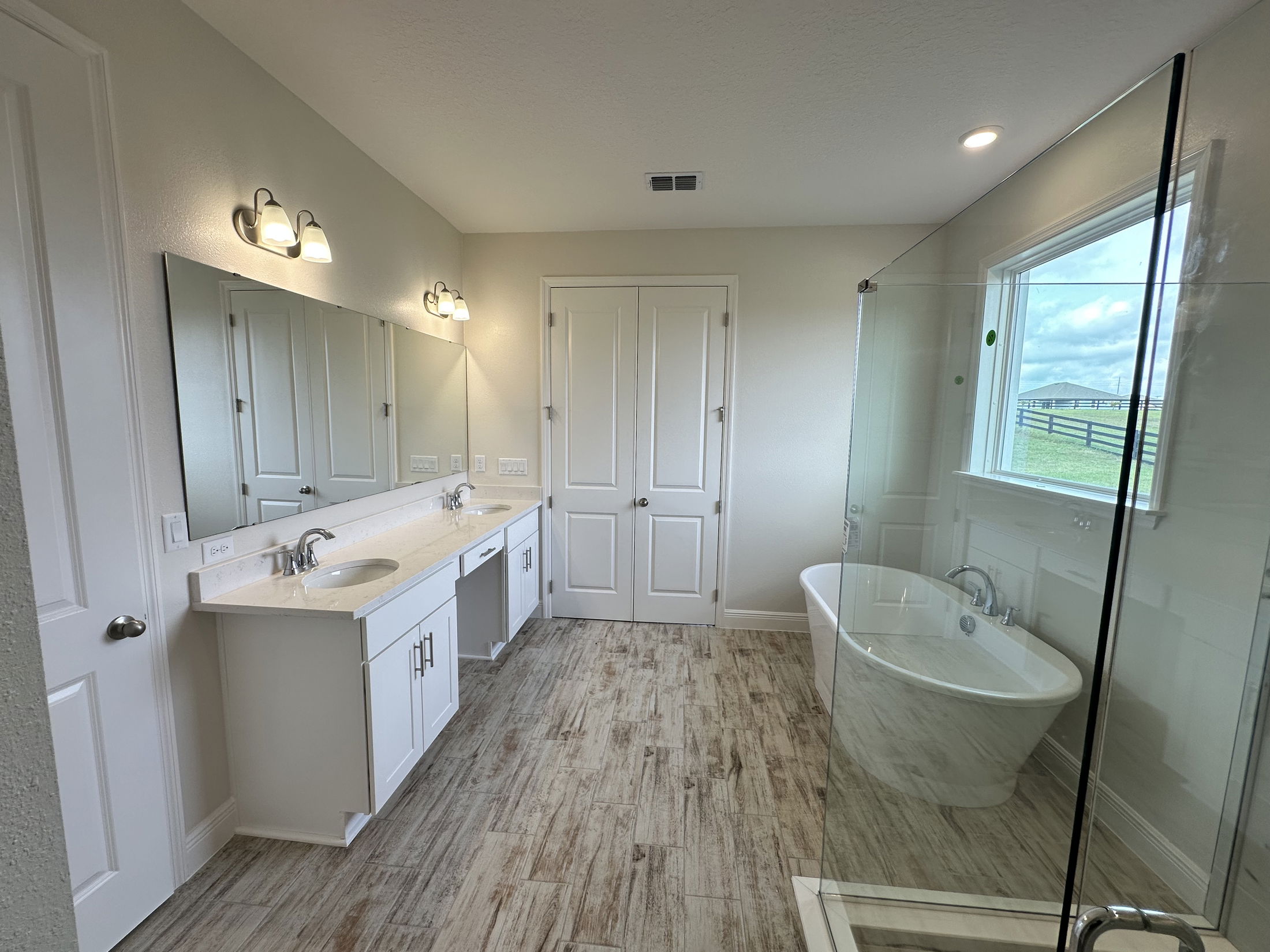 Modern bathroom with double vanity, freestanding bathtub, and glass shower enclosure featuring wood-style flooring and large window.