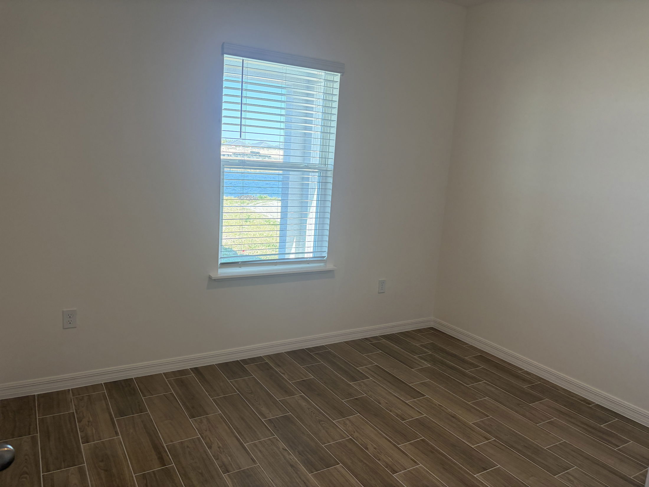 A bright, empty room featuring a large window with blinds and wooden tile flooring, ideal for showcasing modern interior design.