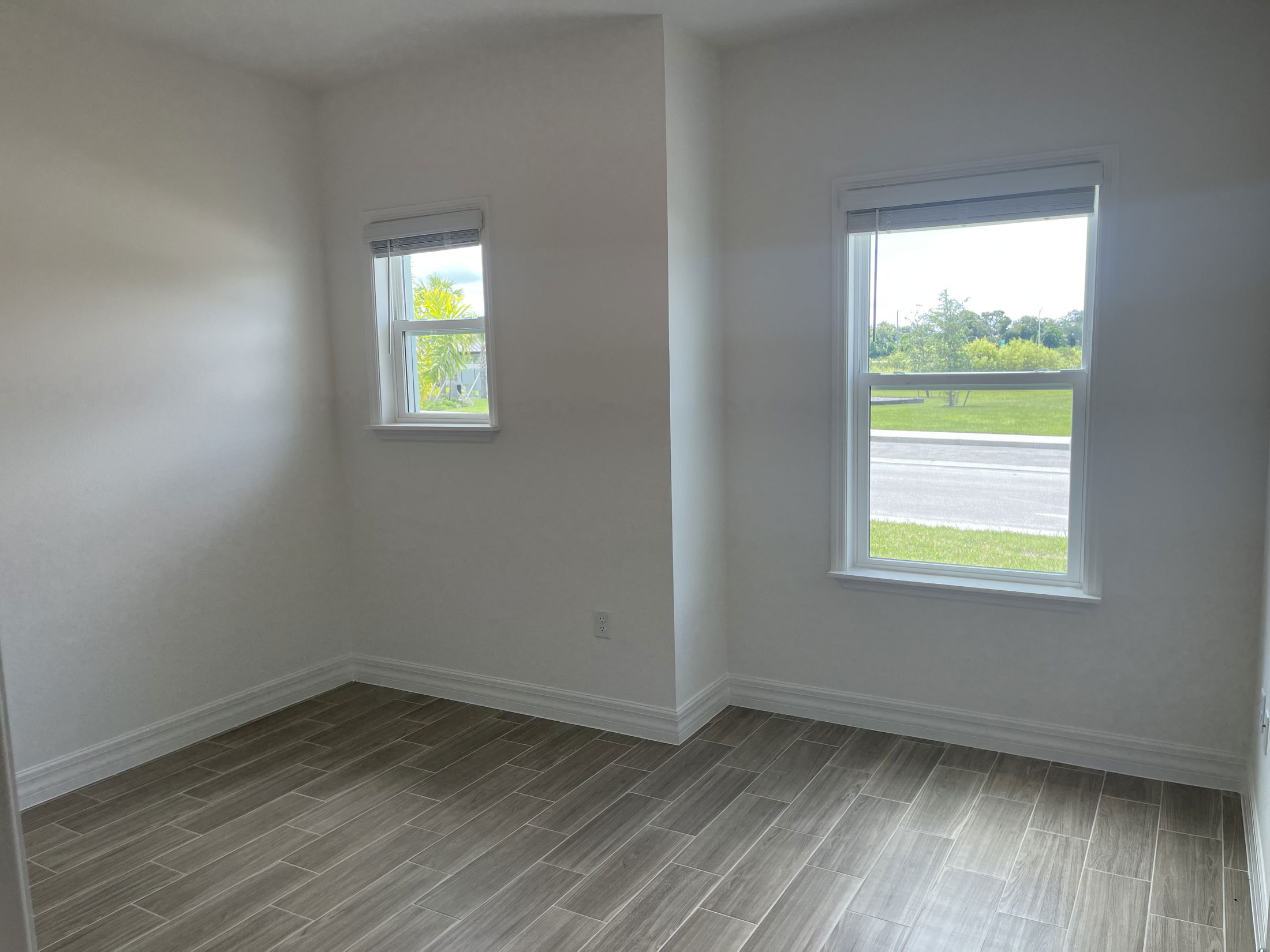 Empty sunlit room with wood-look tiled flooring and two windows overlooking a green yard.