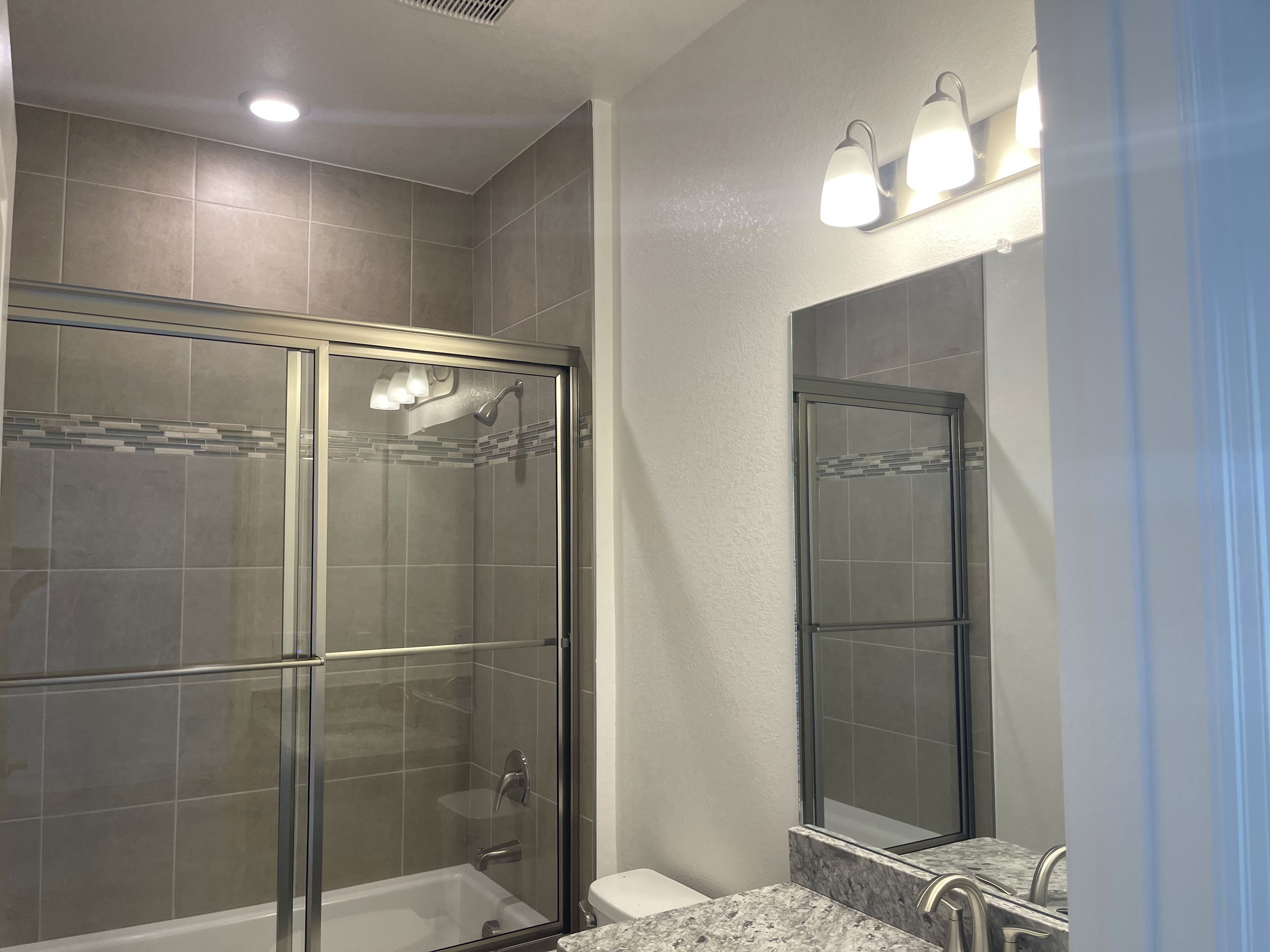 Modern bathroom with a glass-enclosed bathtub, gray tiled walls, and vanity lights above a mirror.