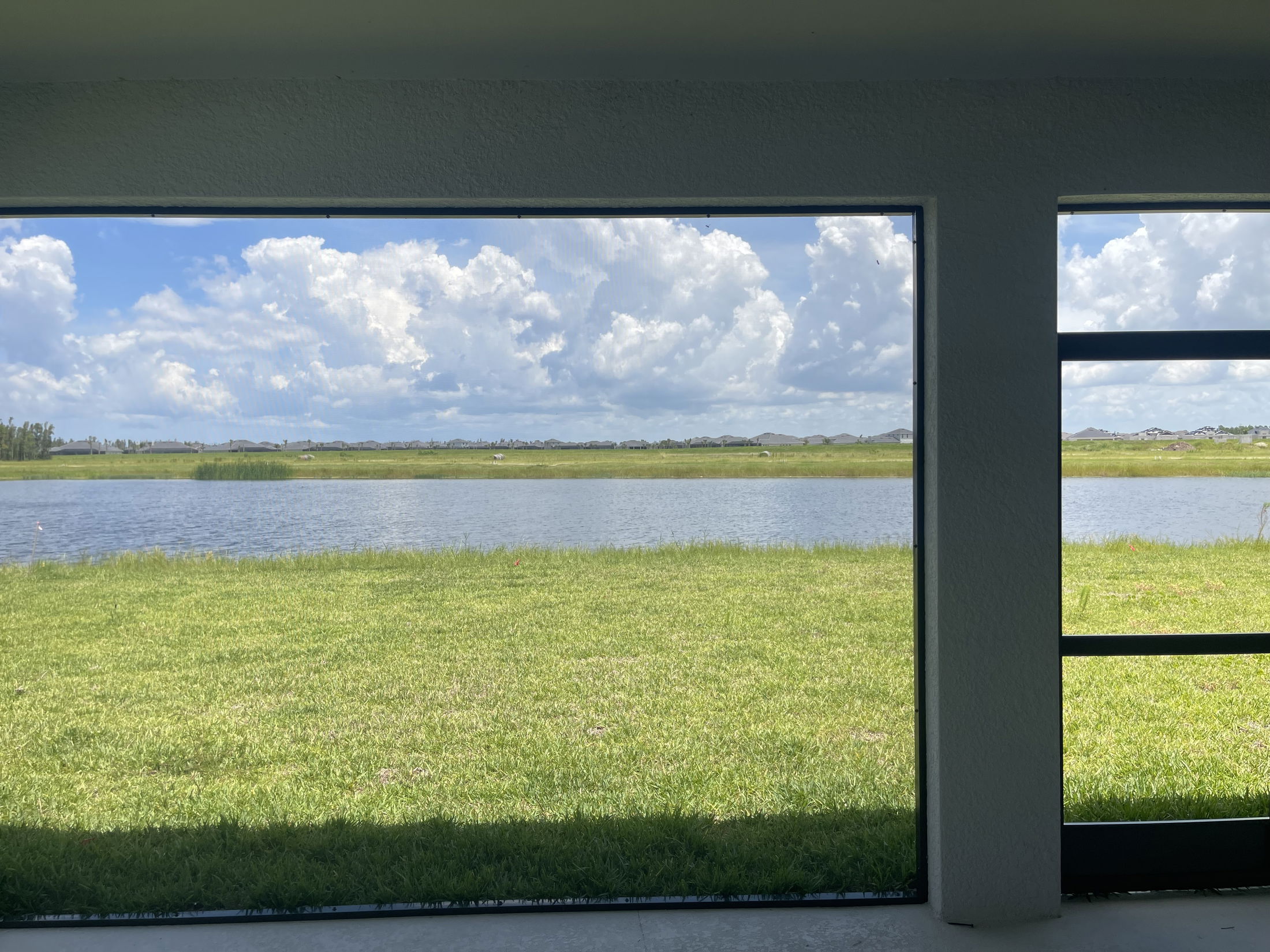 View of a serene lake and grassy field through a large window on a sunny day, with fluffy clouds in the blue sky.
