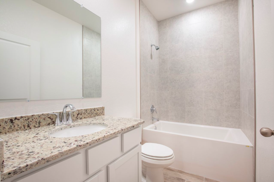 Modern bathroom with granite countertop, white vanity, and bathtub surrounded by gray tile walls.