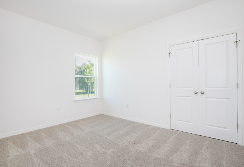 Spacious, well-lit bedroom with beige carpeting and white walls featuring a window and double doors.