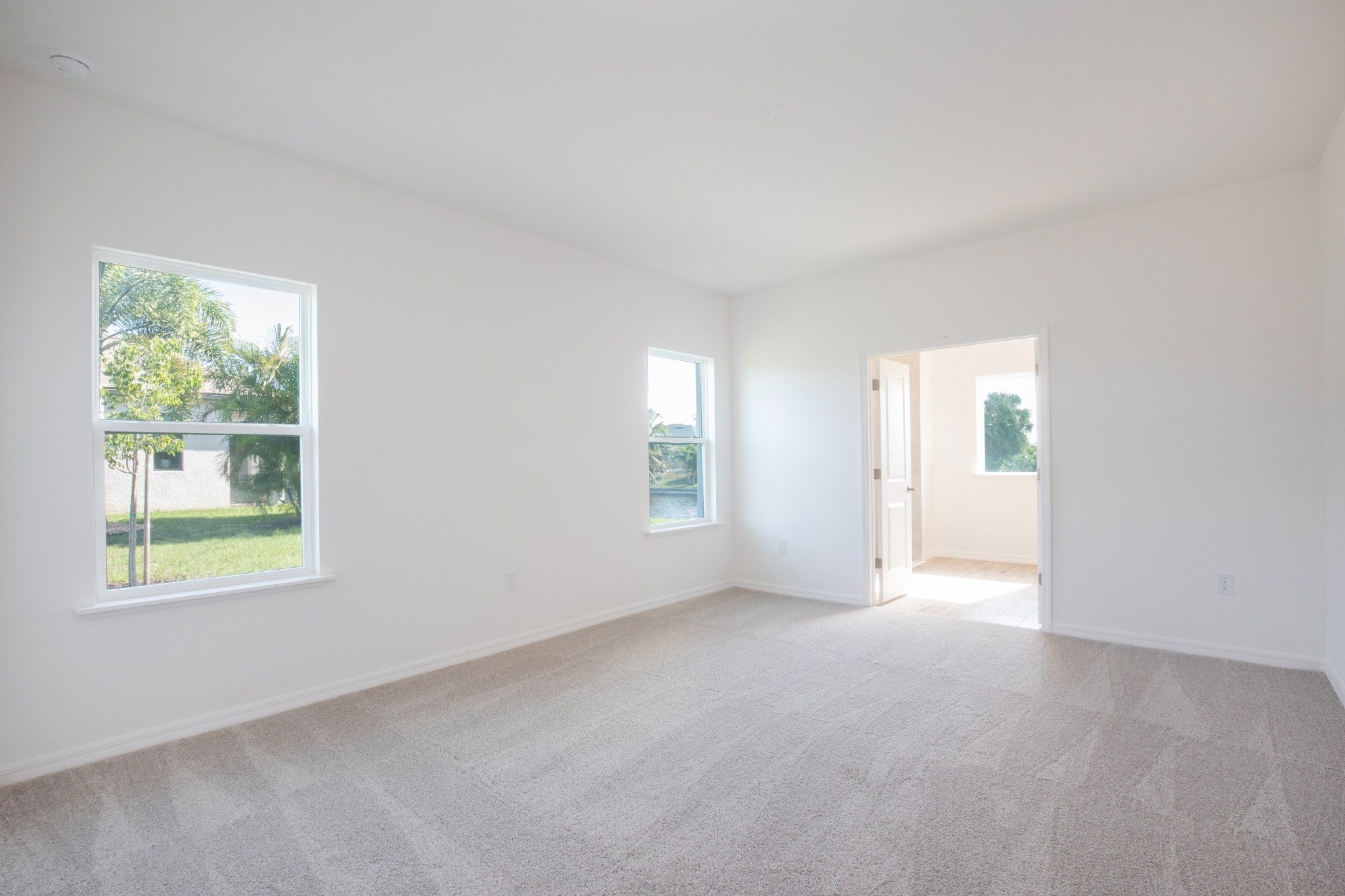 Bright, empty room with beige carpet, white walls, and large windows letting in natural light.