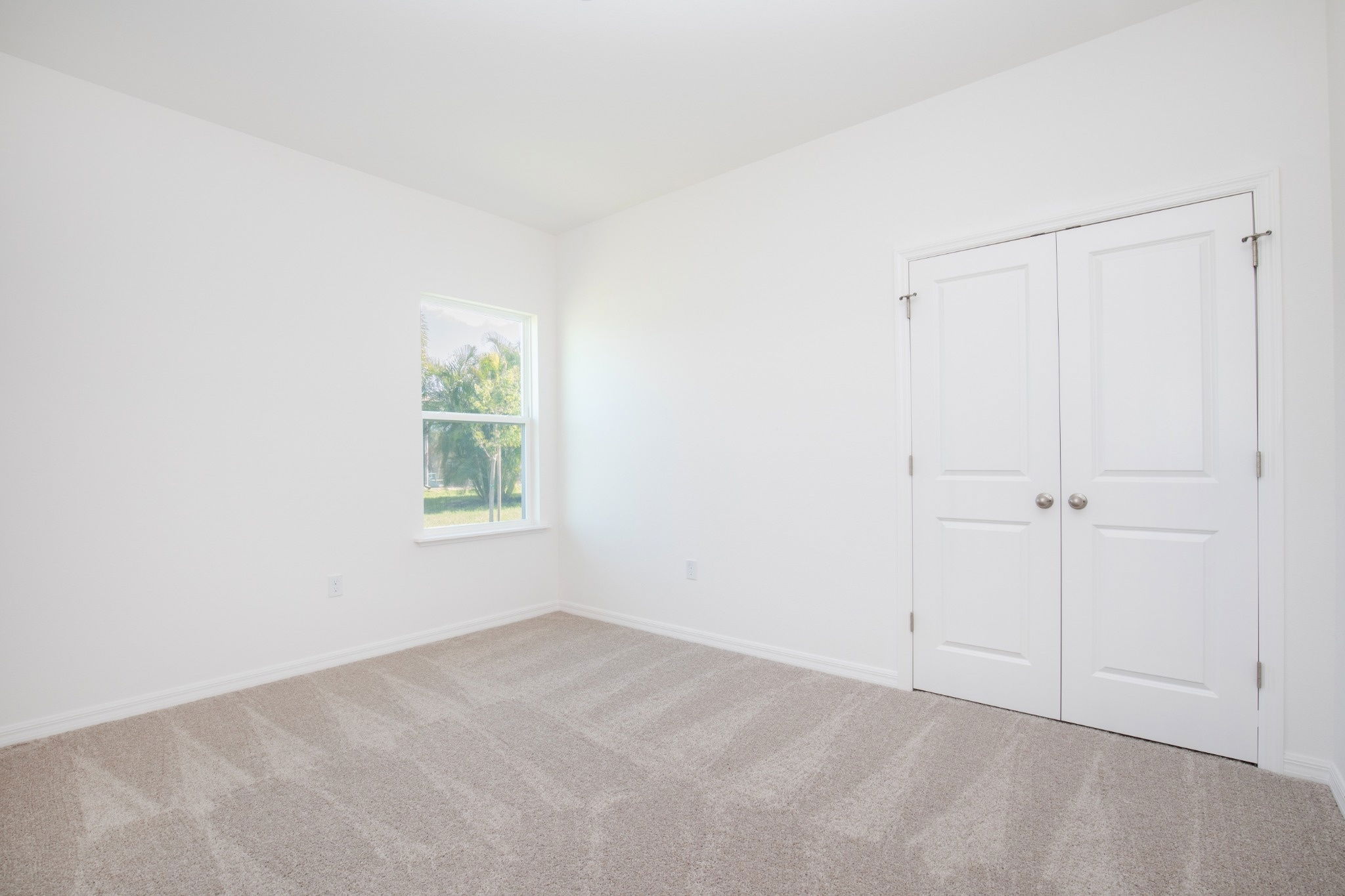 Bright and spacious empty room with beige carpet and white walls, featuring a window and double doors.