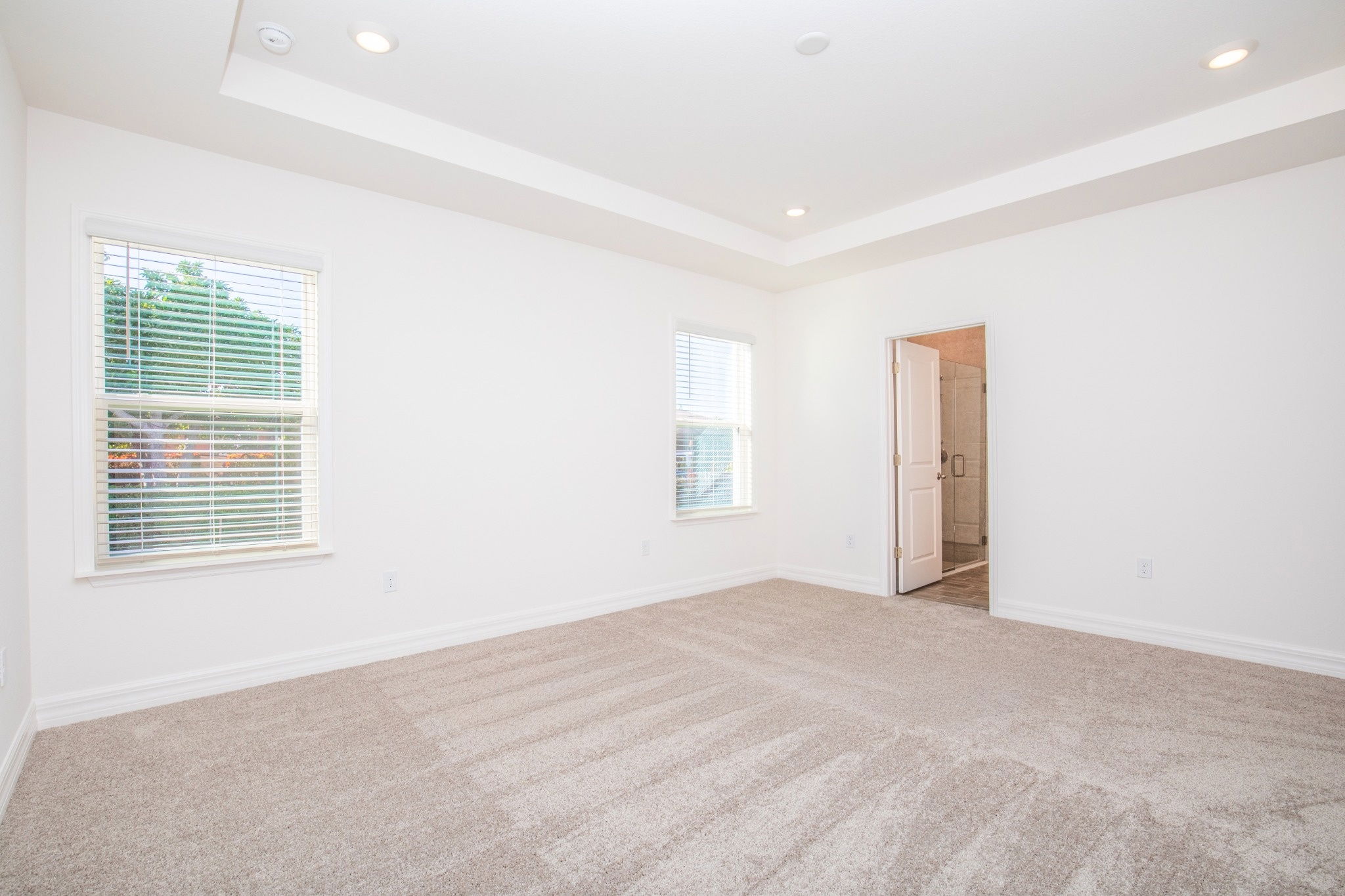Bright and spacious empty bedroom with beige carpet, large windows, and a doorway leading to a bathroom.