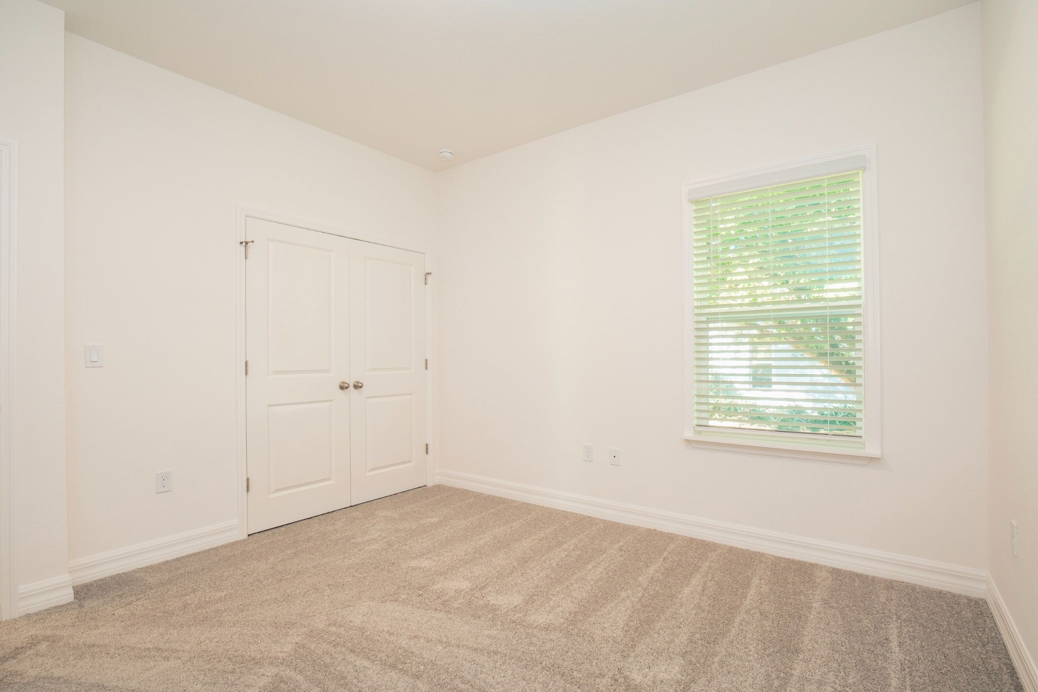 Empty interior room featuring beige carpet, white walls, double closet doors, and a window with blinds.