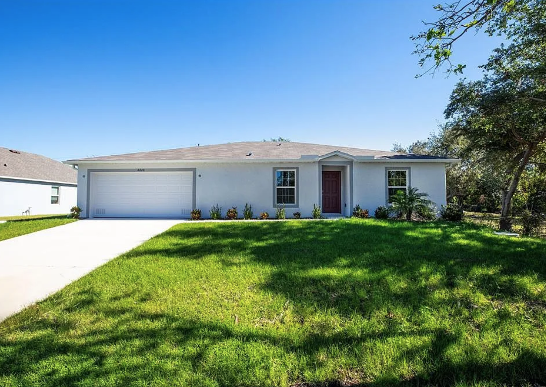 Single-story white house with a two-car garage, red front door, and well-maintained green lawn under a clear blue sky.