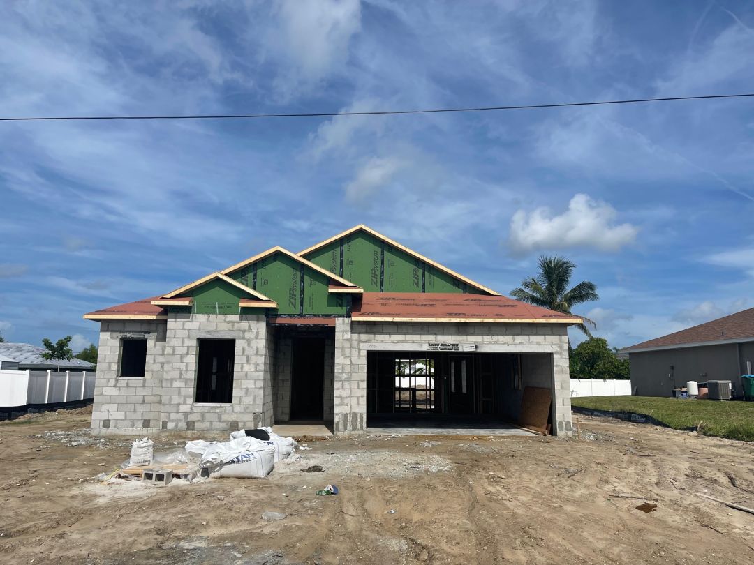 A partially constructed single-story house with exposed brick walls and green roof sheathing under a clear blue sky.