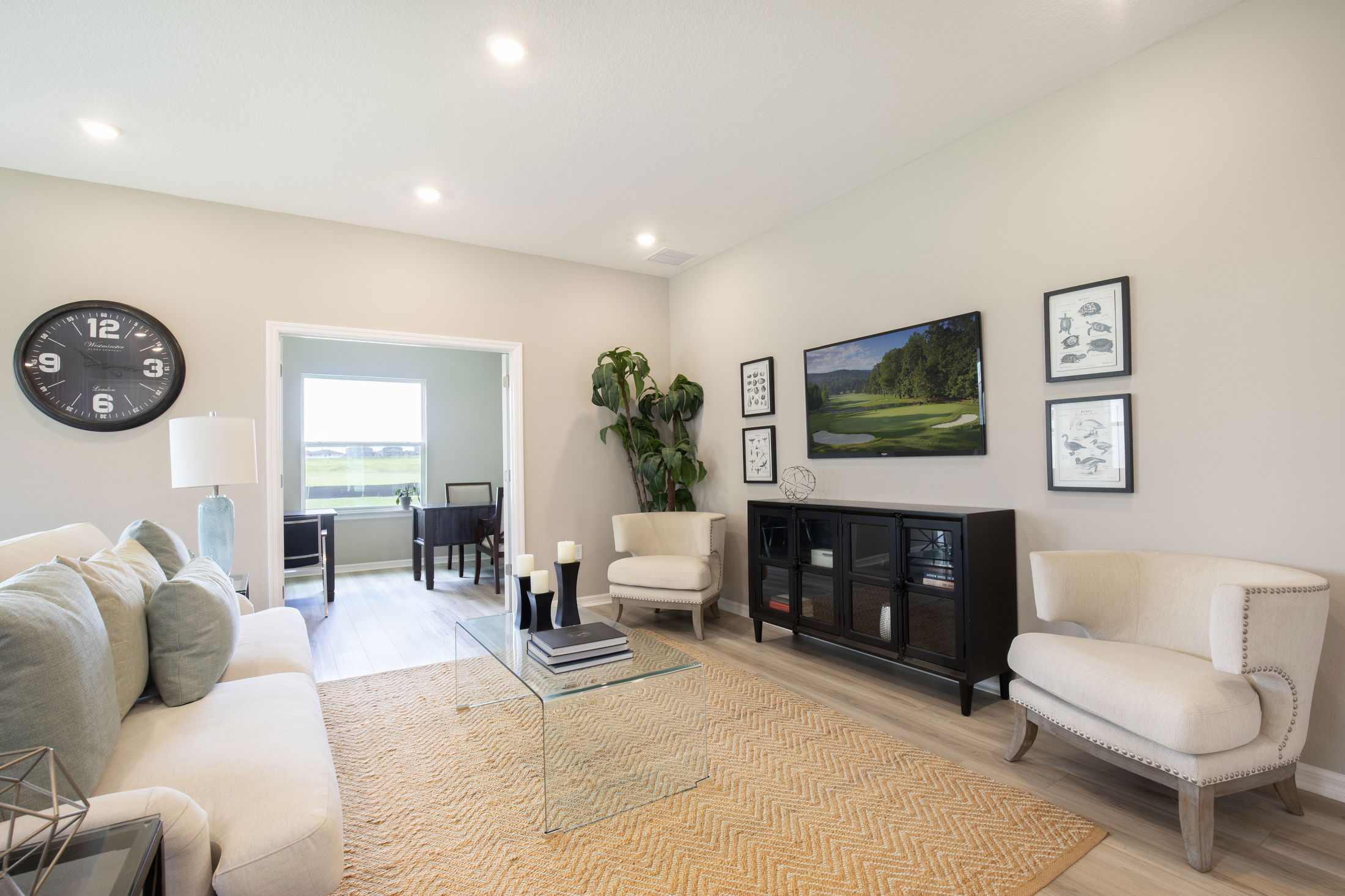Modern living room with beige walls, contemporary furniture, large wall clock, and artwork, leading to a bright study area through white double doors.