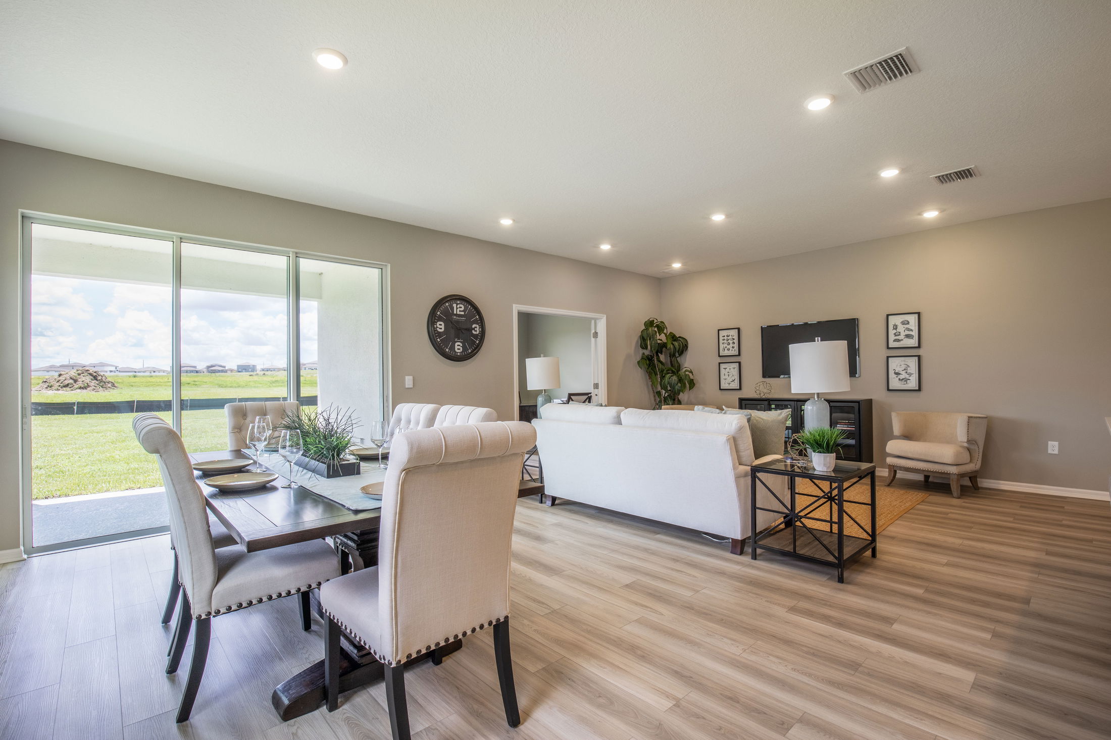 Spacious modern living and dining room interior with neutral tones, wooden flooring, and large windows overlooking a green lawn.