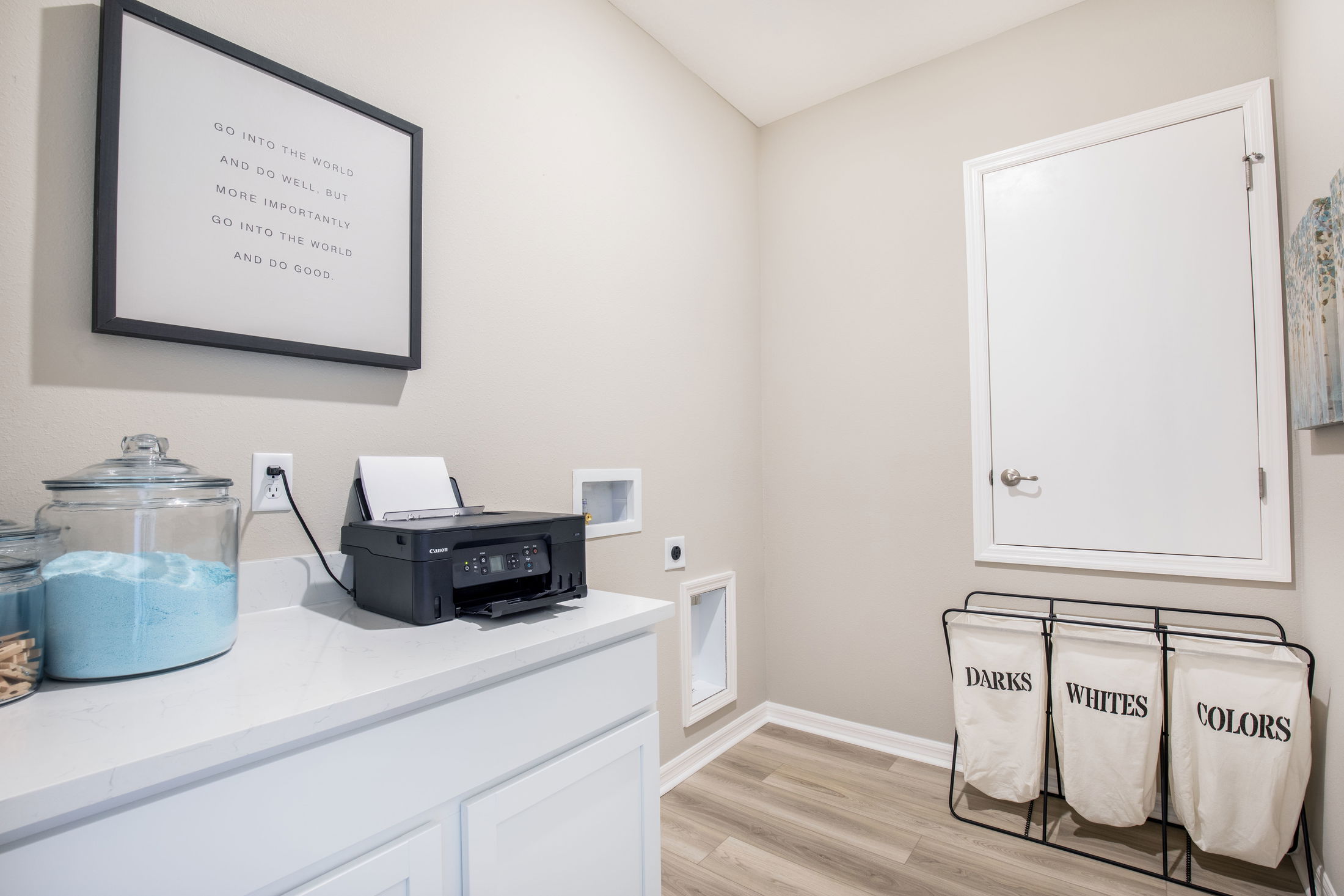 Spacious laundry room with a printer, detergent jars, inspirational wall art, and a three-compartment laundry sorter on light wood flooring.