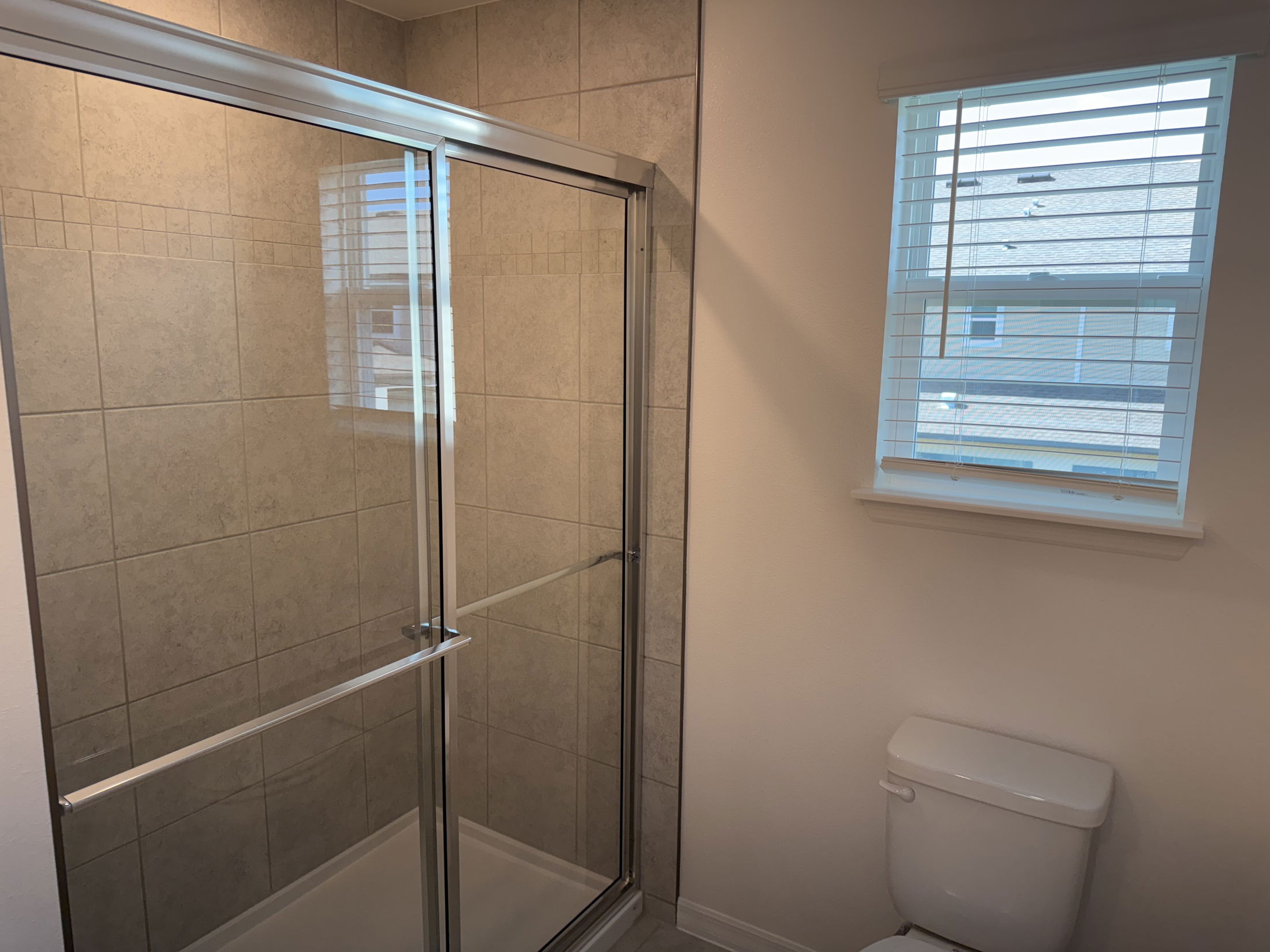Modern bathroom with beige tiled shower and glass doors next to a window with blinds.