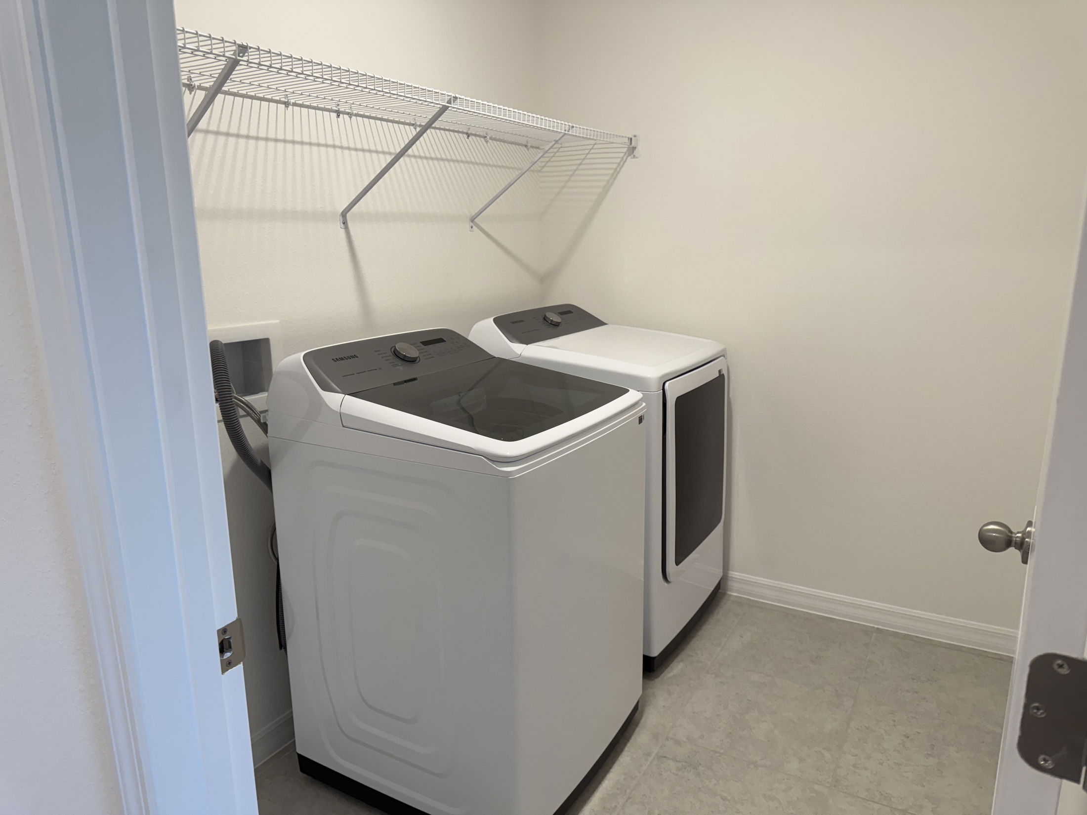 Laundry room with Samsung washer and dryer, featuring tiled floor and wall-mounted wire shelf.