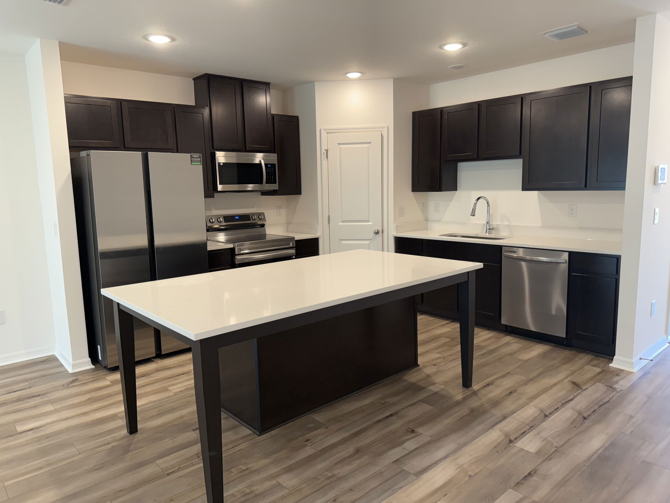 Modern kitchen with dark wood cabinets, stainless steel appliances, and a large central island on wood-look flooring.