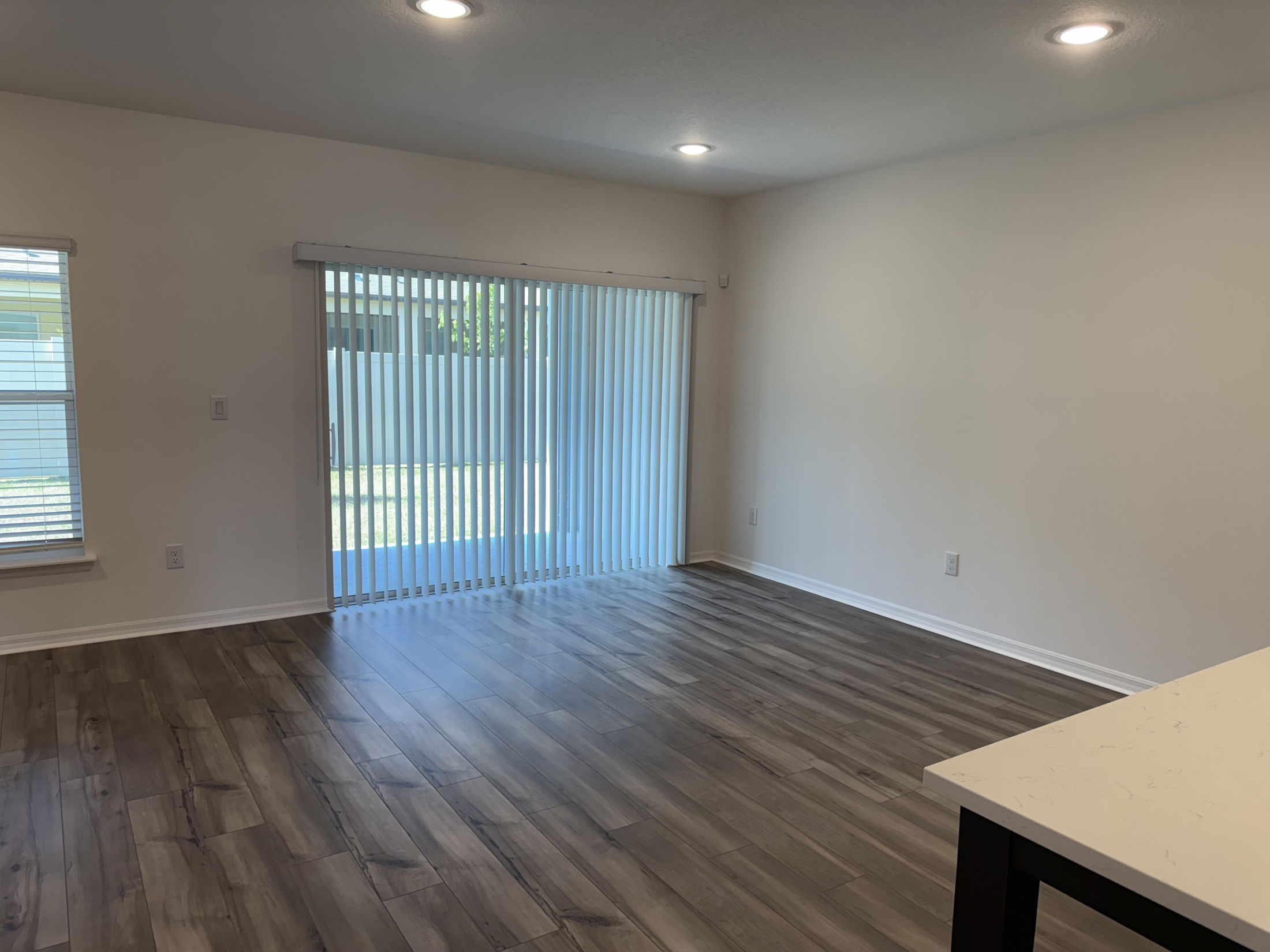 Spacious empty living room with wooden flooring, sliding glass doors, and vertical blinds.