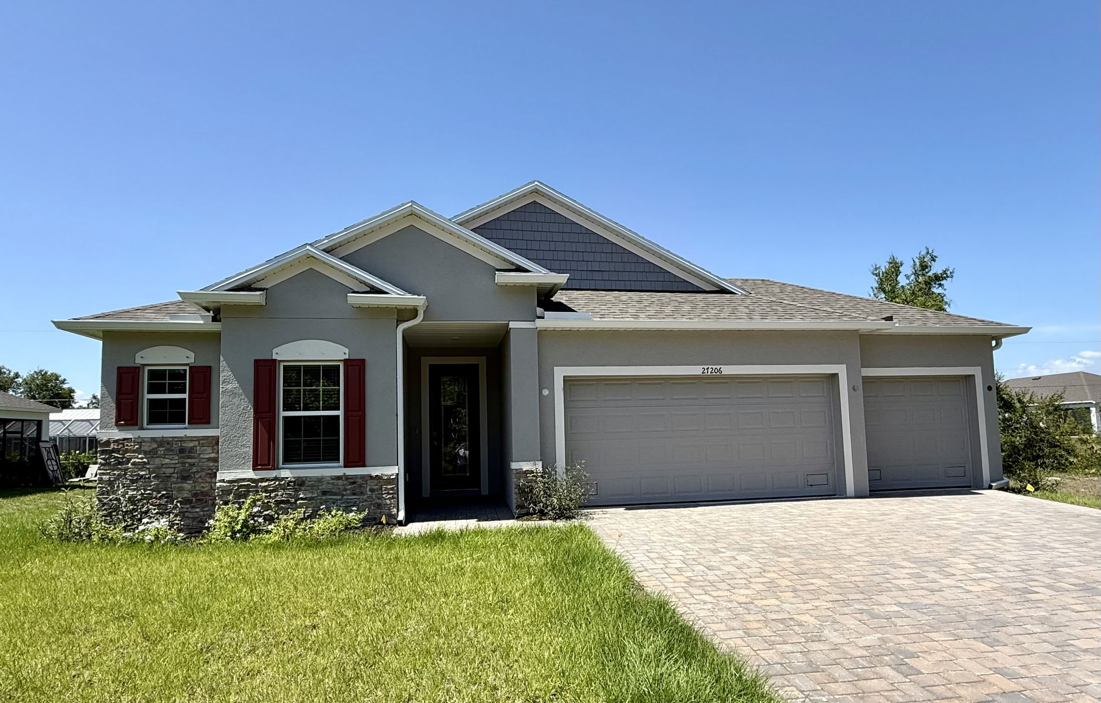 Modern single-story gray house with red shutters, stone accents, and a three-car garage under a clear blue sky.