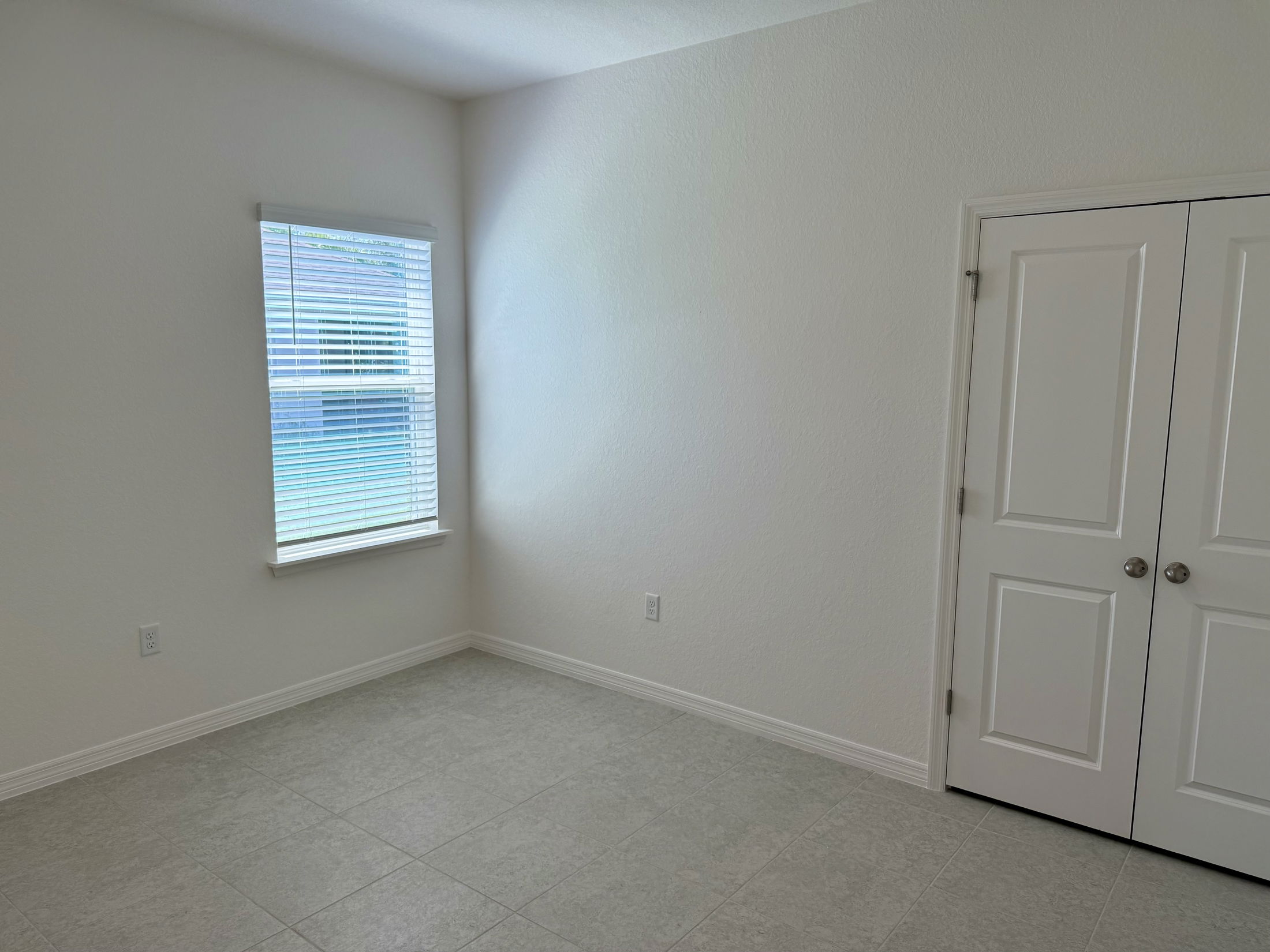 Bright empty room with white walls, tiled floor, a window with blinds, and a closed closet door.