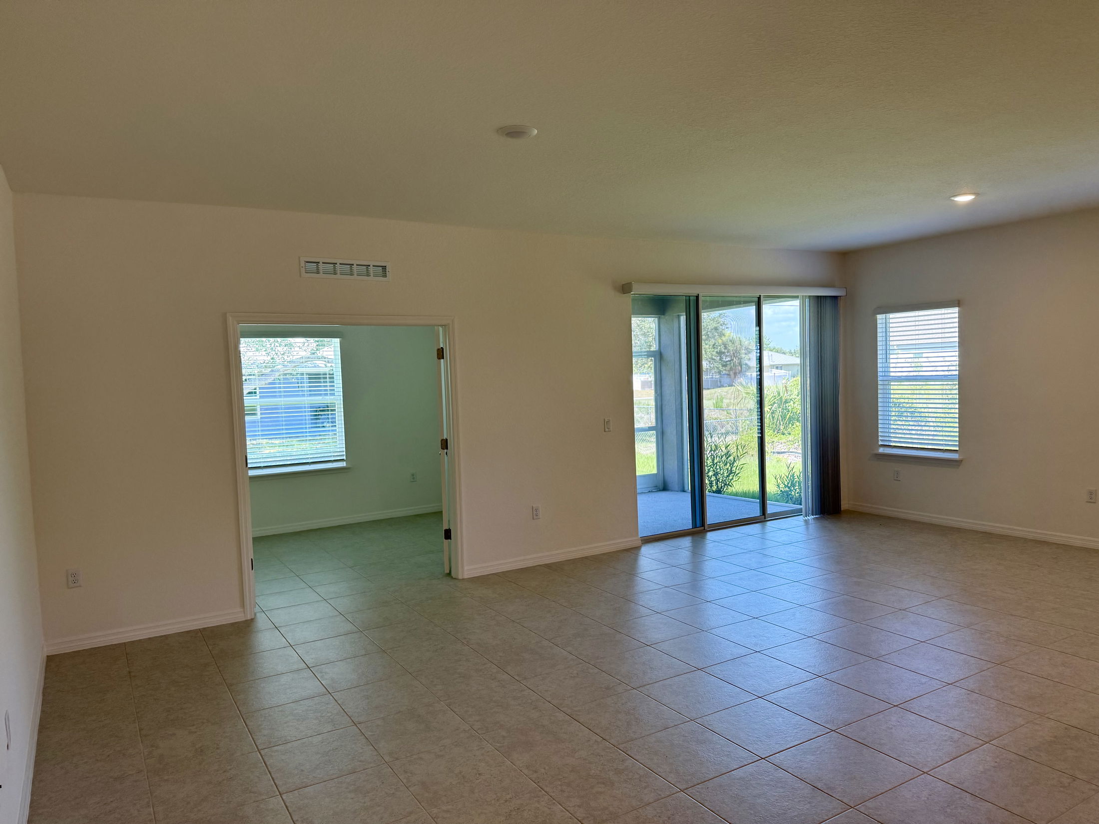 Bright, empty living room with tiled floor, sliding glass doors to a garden, and a view into an adjoining room.