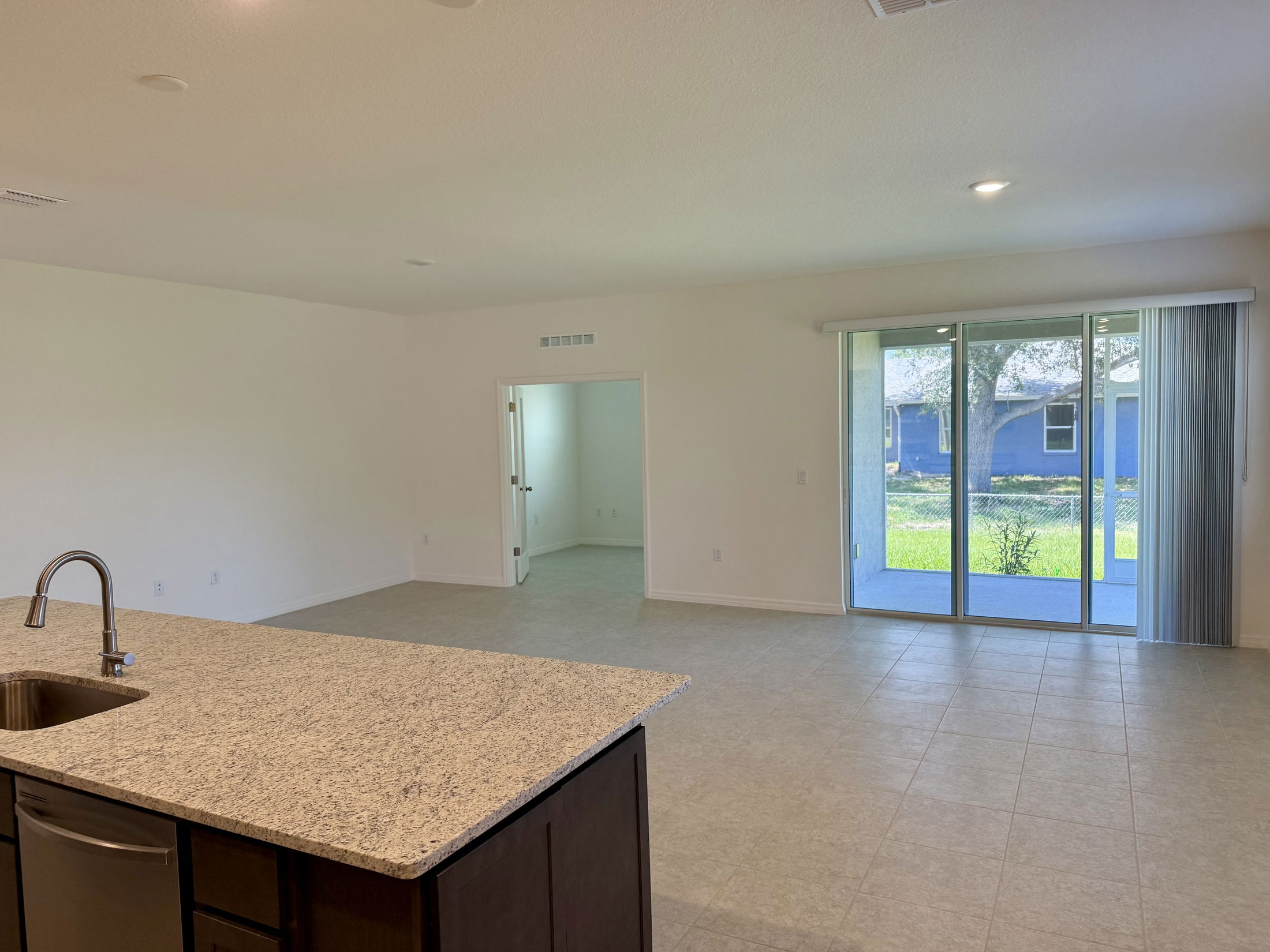 Open-concept living room with large glass sliding doors, granite kitchen countertop, and tile flooring, leading to a bright backyard view.