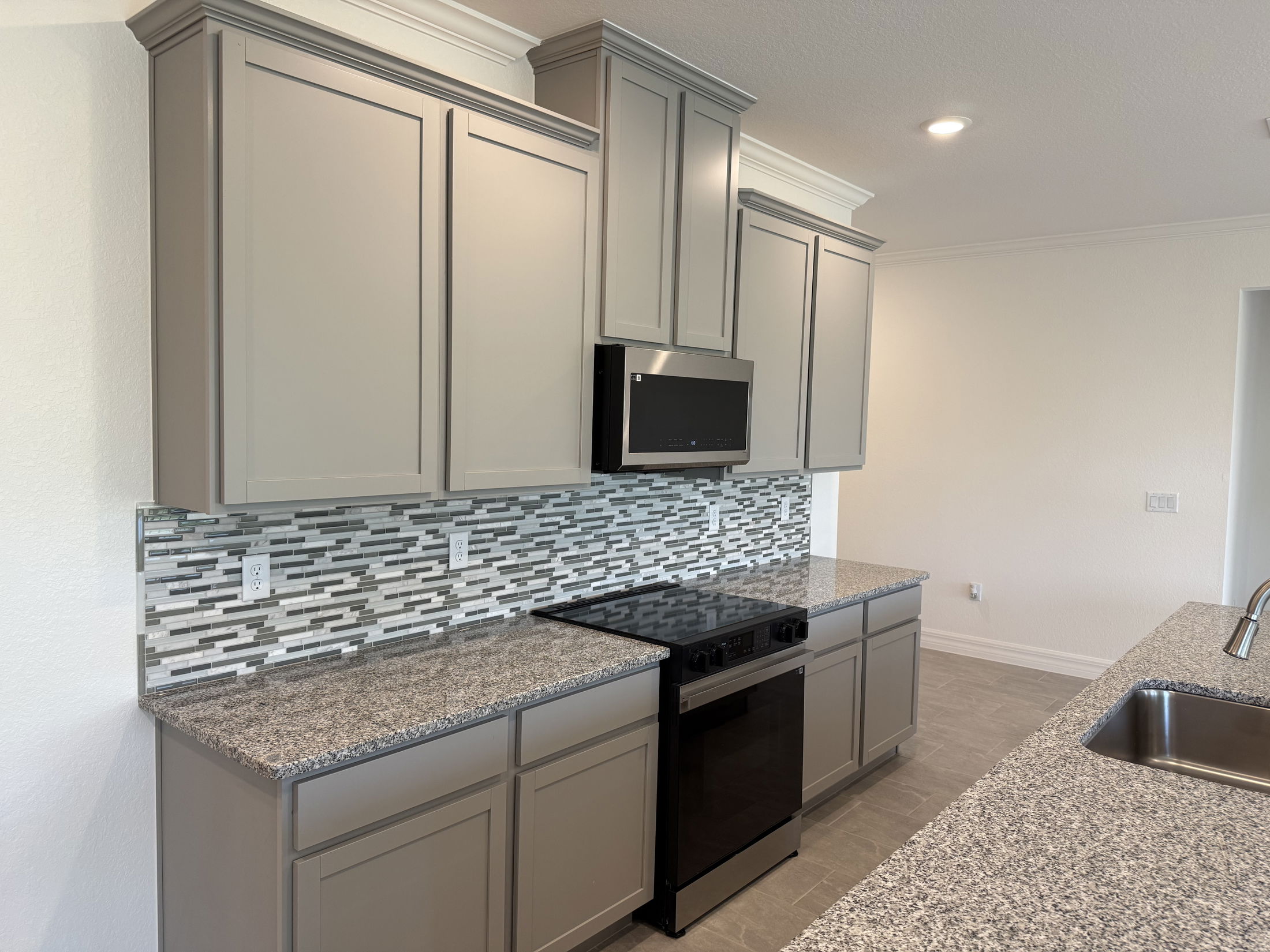 Modern kitchen with gray cabinets, mosaic tile backsplash, granite countertops, black appliances, and recessed lighting.