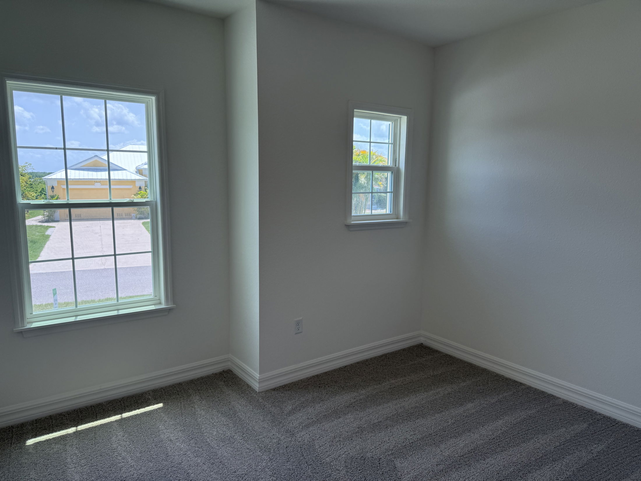 Bright empty room with large windows, soft carpet, and a clear view of the outside street and house.