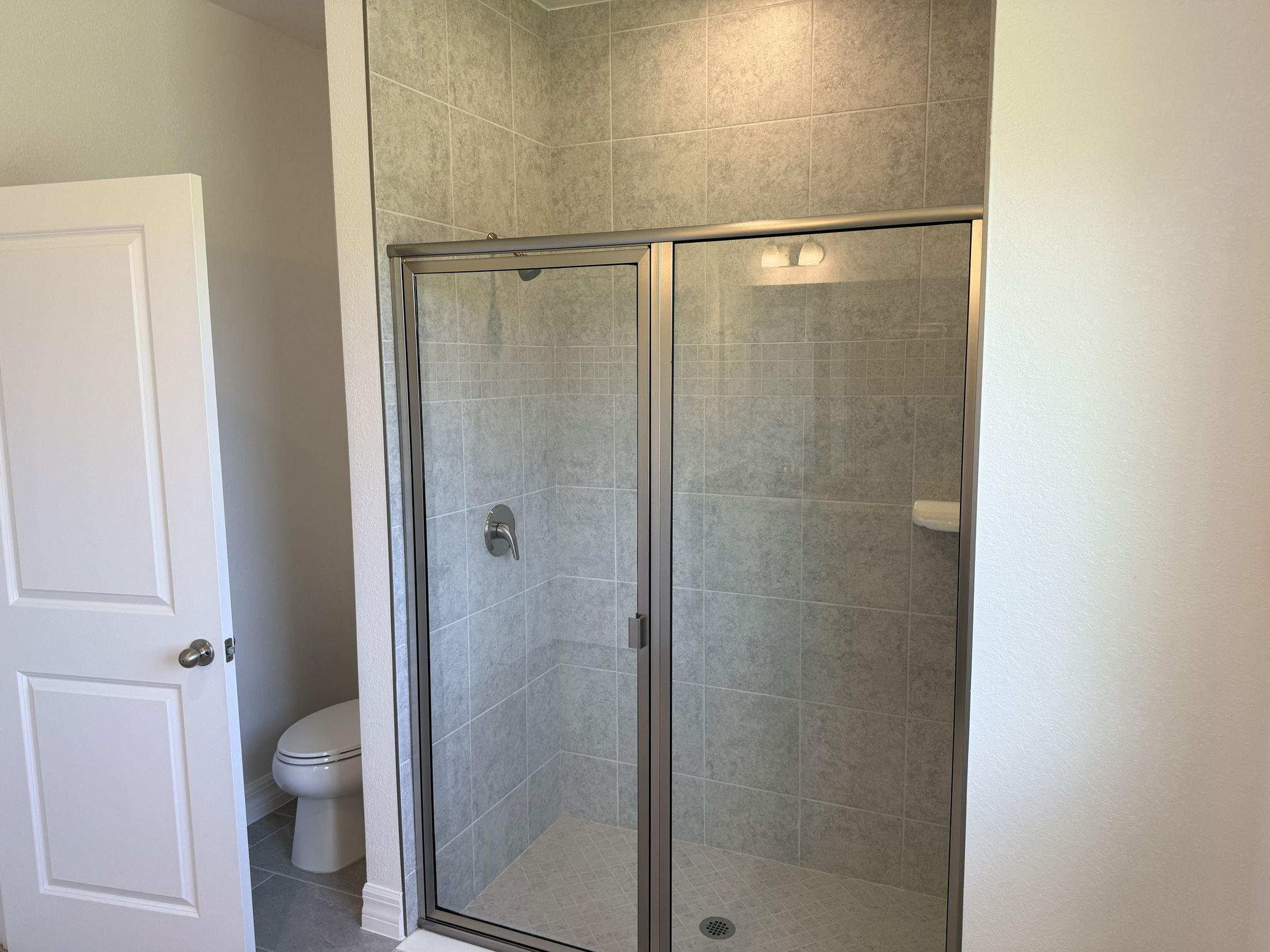 Modern bathroom featuring a glass-enclosed shower with gray tile walls and a visible open door leading to a toilet area.