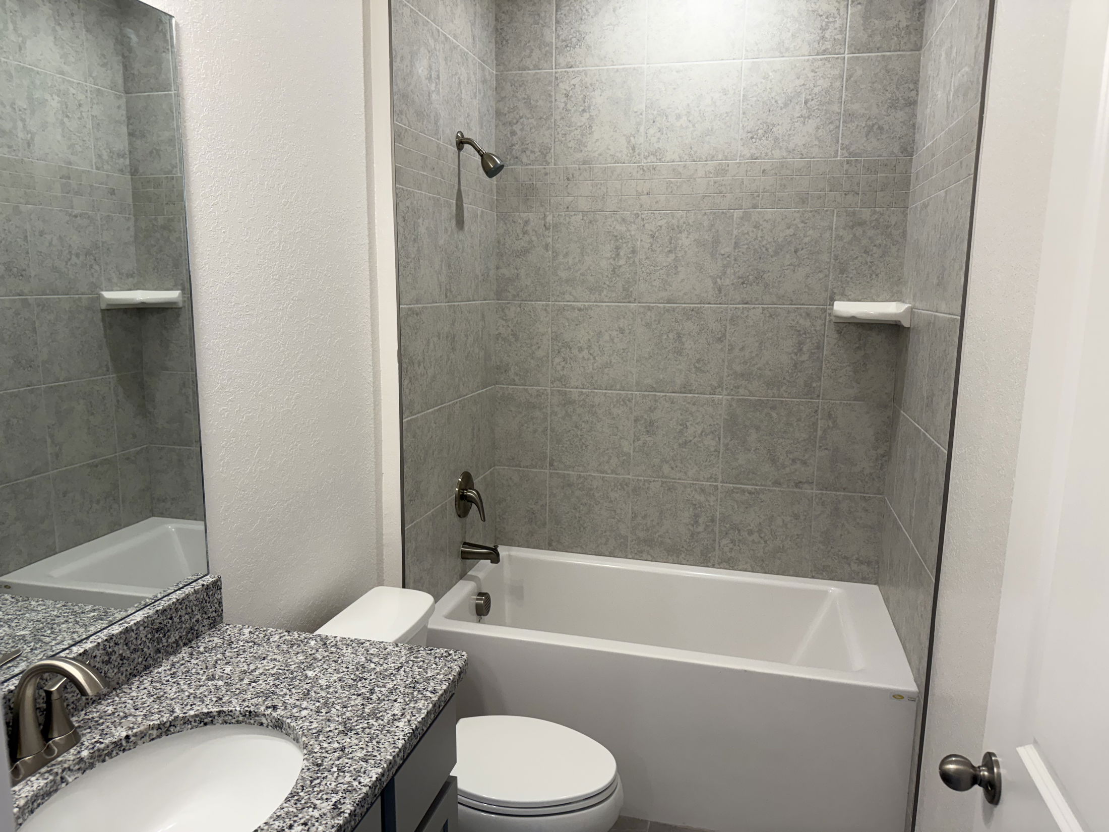 Modern bathroom interior featuring a granite countertop sink, toilet, and bathtub with gray tiled walls.